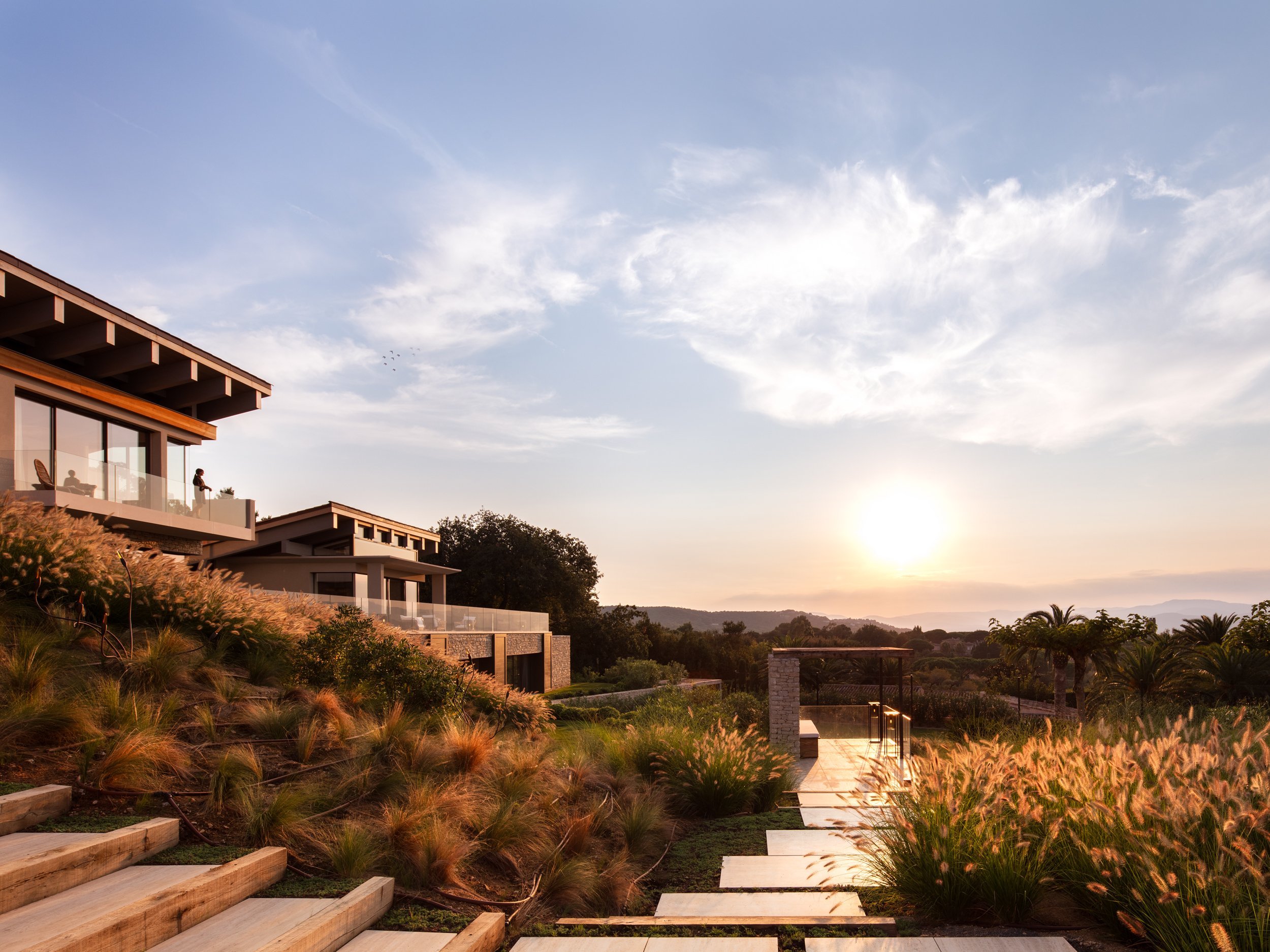 Modern house with large glass windows on a hill at sunset, surrounded by landscaped garden with plants and trees, and a pathway leading to the house.