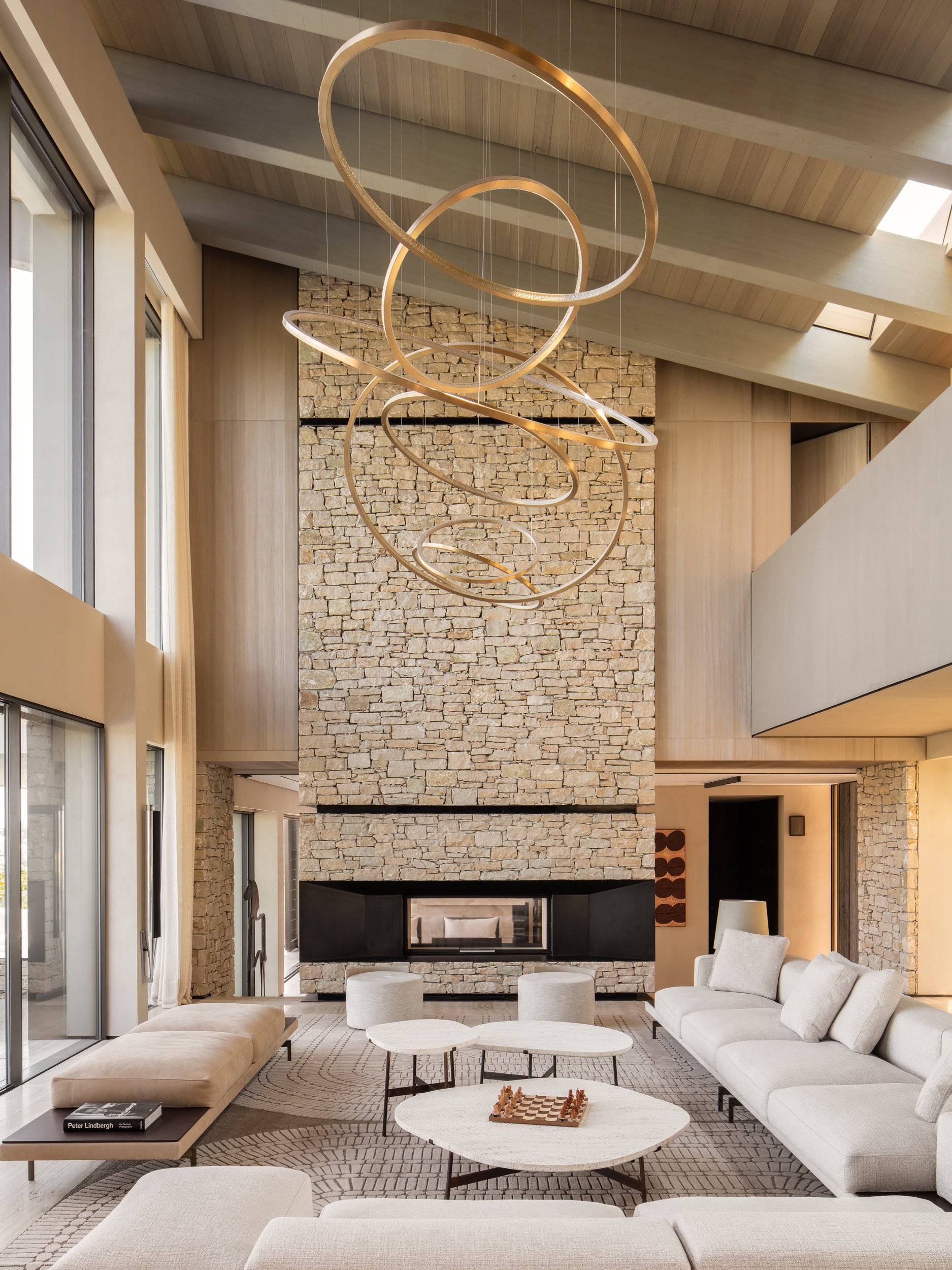 Modern living room with stone fireplace, white sectional sofa, matching ottomans, coffee tables, contemporary chandelier, large windows, and wooden ceiling beams.