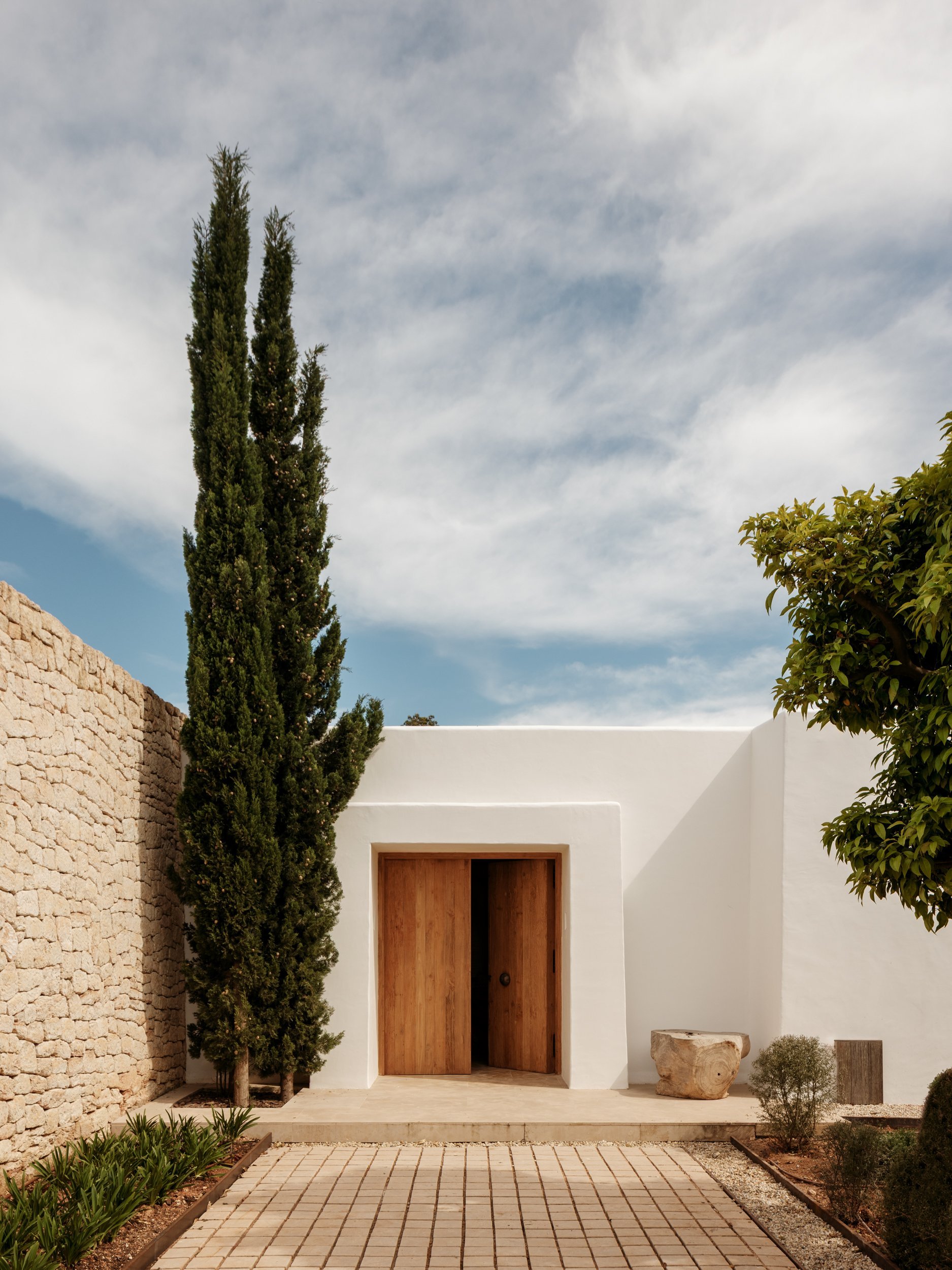 Modern white house with wooden door, tall cypress tree, small shrub, and stone decorations in a landscaped garden under a cloudy sky.
