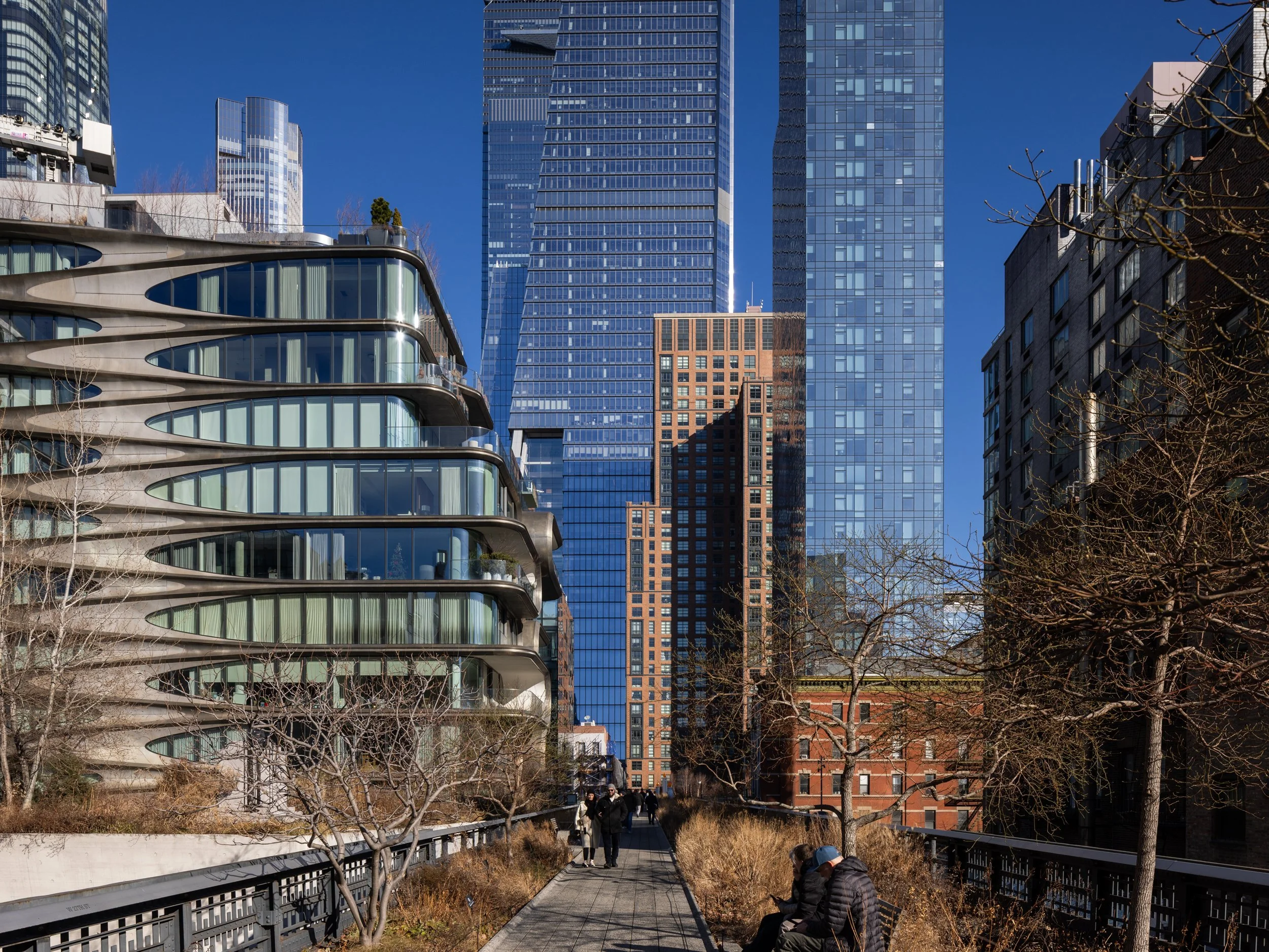 Urban cityscape with modern high-rise buildings and a unique curved glass building, a pedestrian walkway, leafless trees, and people sitting on benches under a clear blue sky.