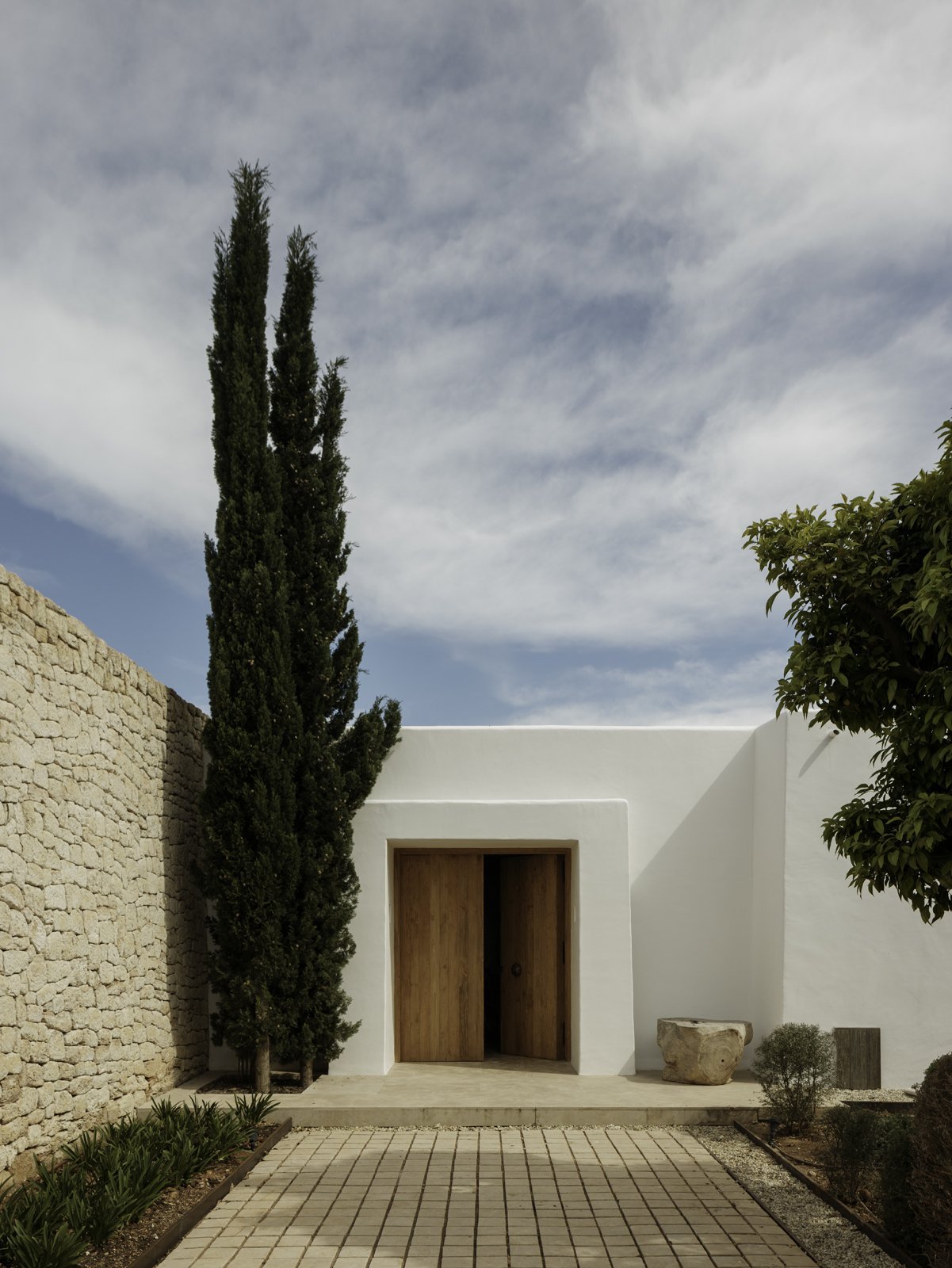A modern white house with a wooden door, tall cypress tree on the left, and landscaped front yard with small shrubs and stone accents.