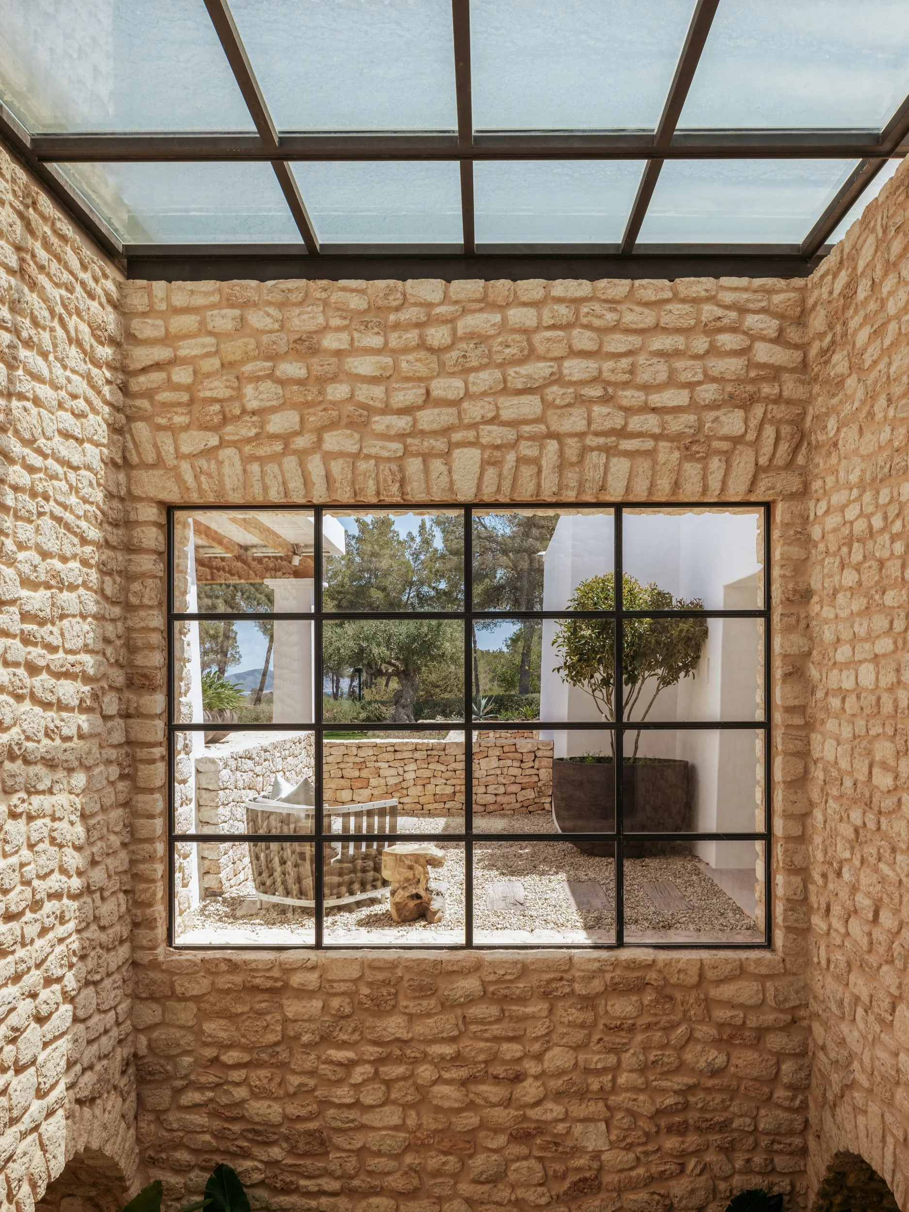Interior view of a stone-walled room looking out through a large multi-pane window to an outdoor patio with a large plant in a planter, a chair, small table, rocks, trees, and a view of the sky and distant scenery.
