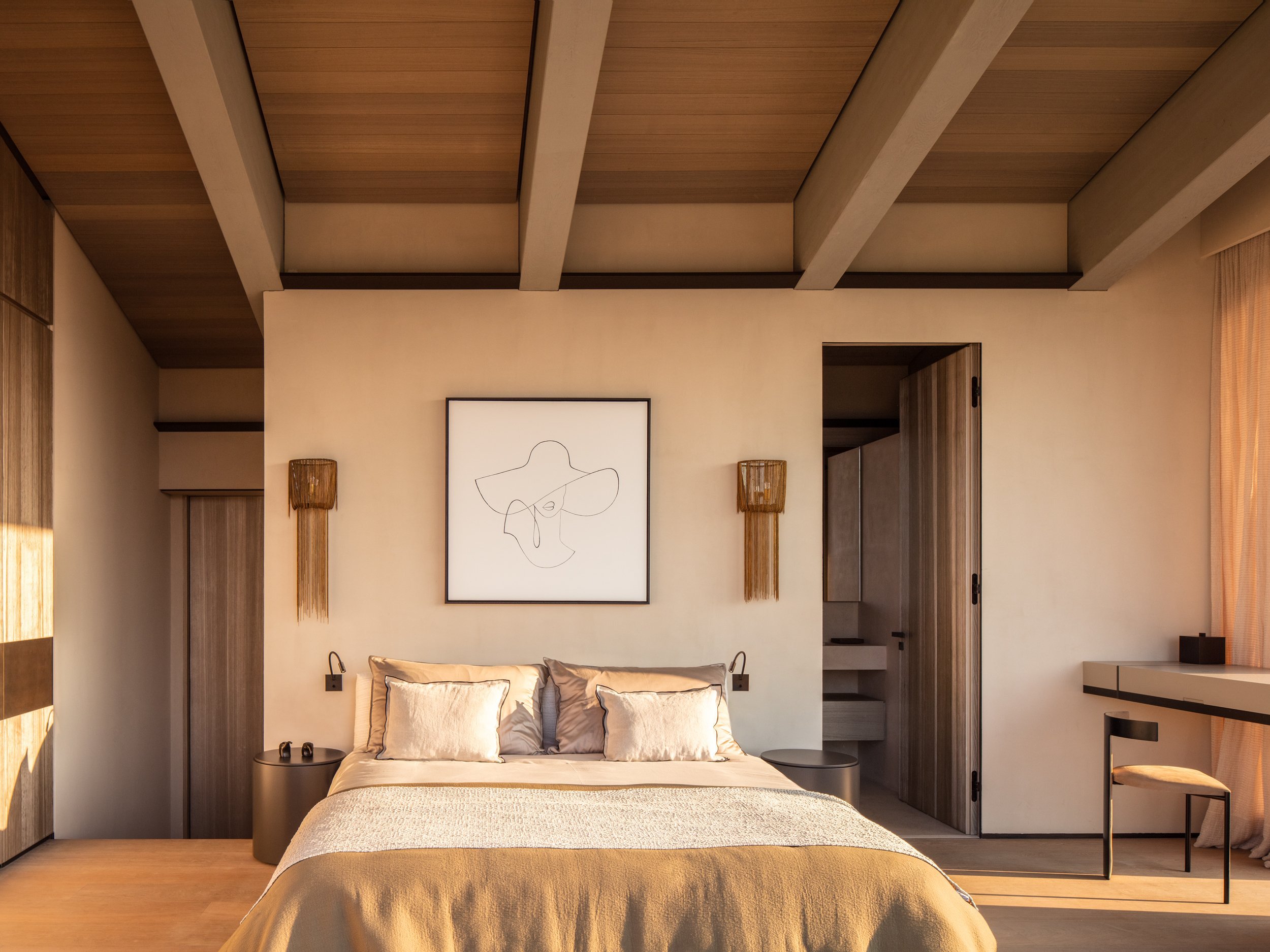 A minimalist bedroom with a large bed, beige bedding, and pillows, framed art above the headboard, wooden wall accents, bedside tables, and a chair near a window with sheer curtains.