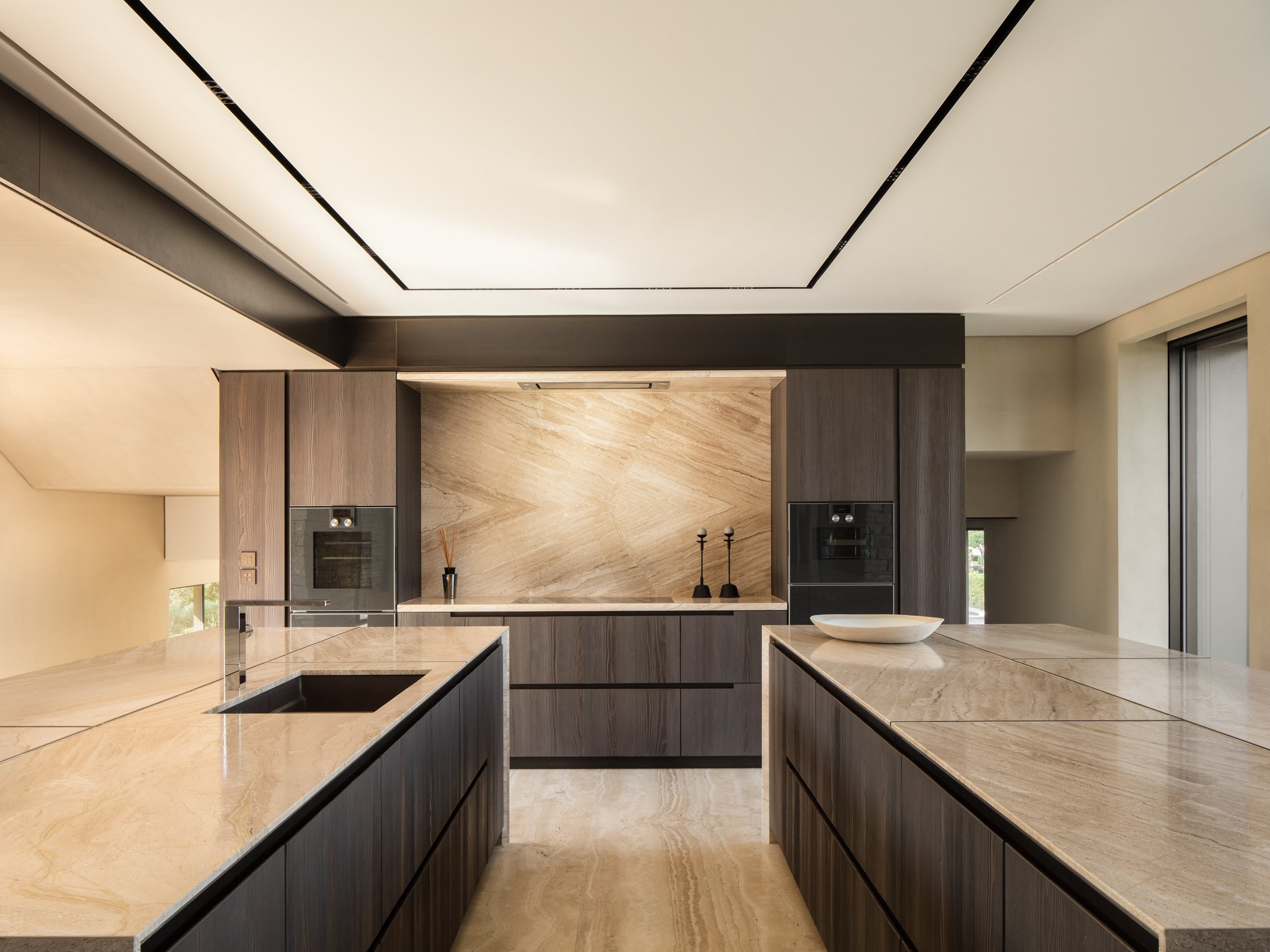 Modern kitchen with beige marble countertops, dark wood cabinets, and a beige marble backsplash. There is a central island with a built-in black sink and a single basin. The ceiling is white with black trim. Large windows provide natural light.