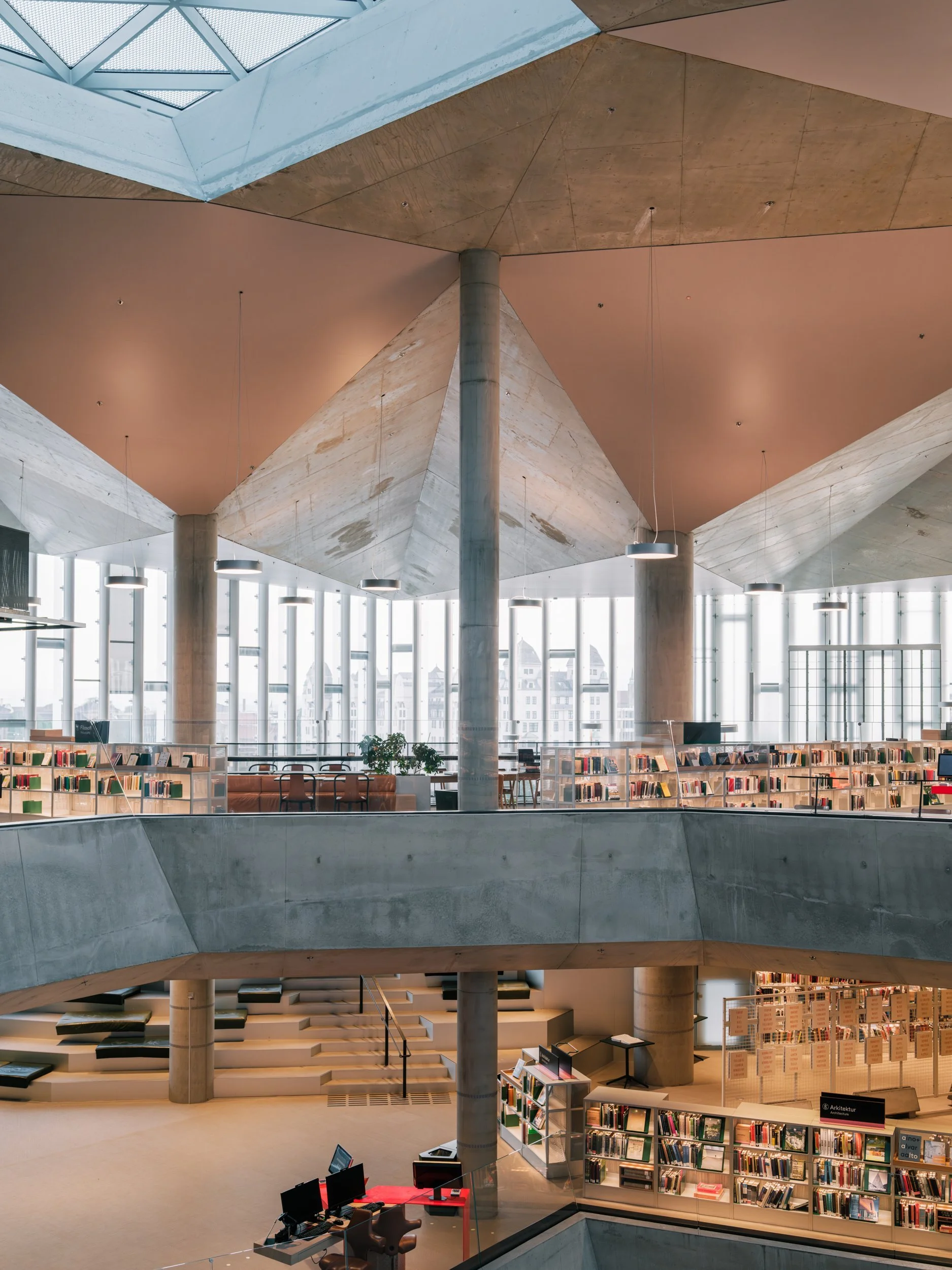 Interior of a modern library with multi-level design, concrete columns, large glass windows, bookshelves, seating area, and staircases.