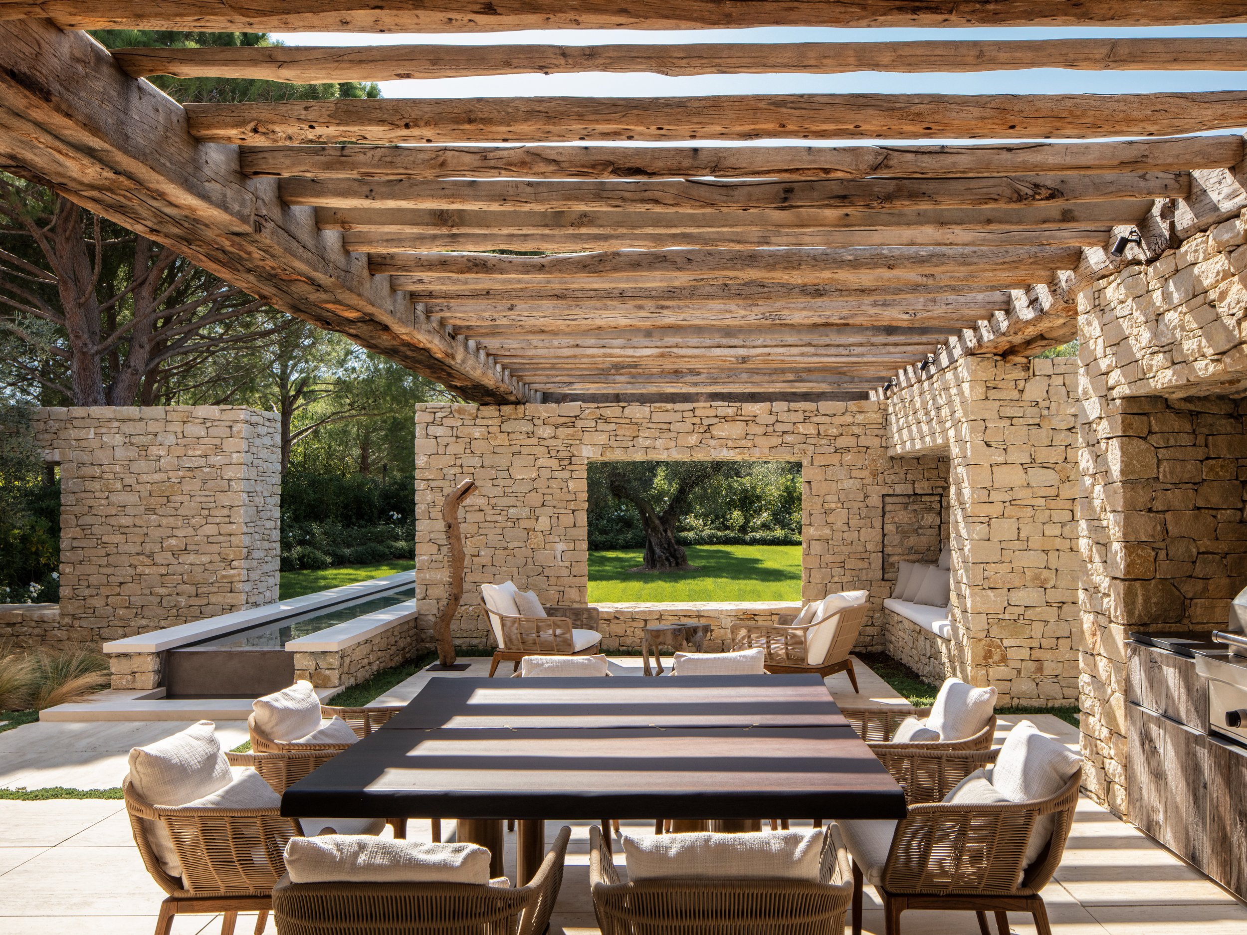 An outdoor patio with a wooden pergola roof, stone walls with large open windows, and modern seating around a black table. The space overlooks a green lawn with trees.