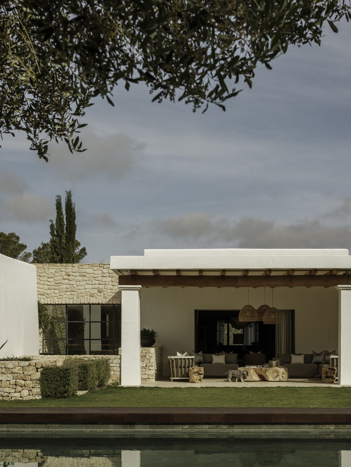 Modern outdoor patio with a sectional sofa, wooden coffee table, and hanging wicker pendant lights, adjacent to a pool under a clear sky.