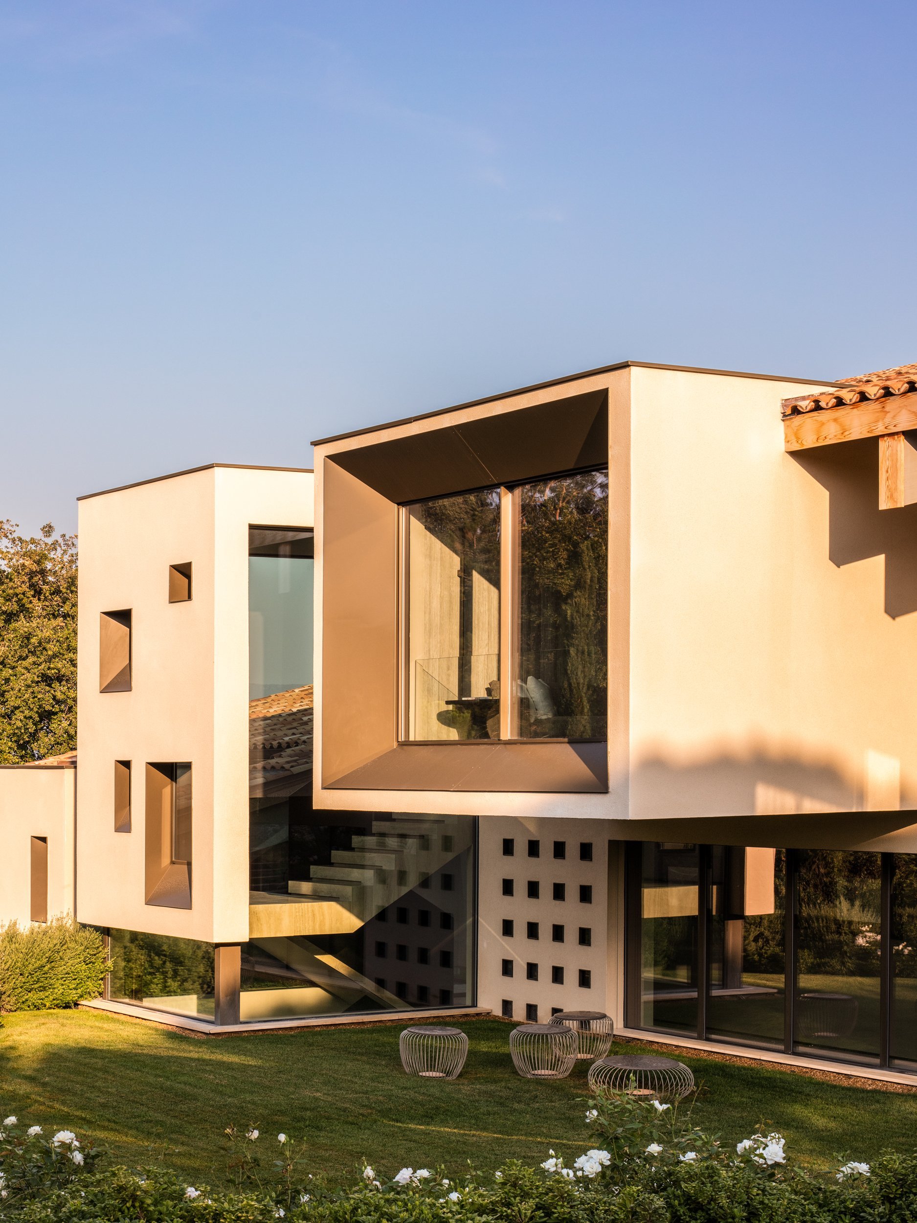 Modern multi-story house with large glass windows, beige exterior walls, and a landscaped front yard with seating.