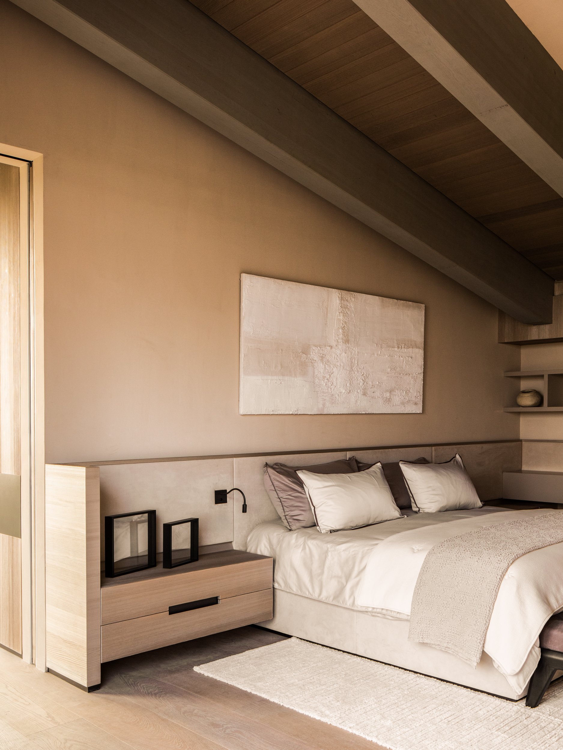 Modern bedroom with beige walls, a large bed with white bedding and multiple pillows, a wooden nightstand with black picture frames, and a textured painting hanging above the bed.