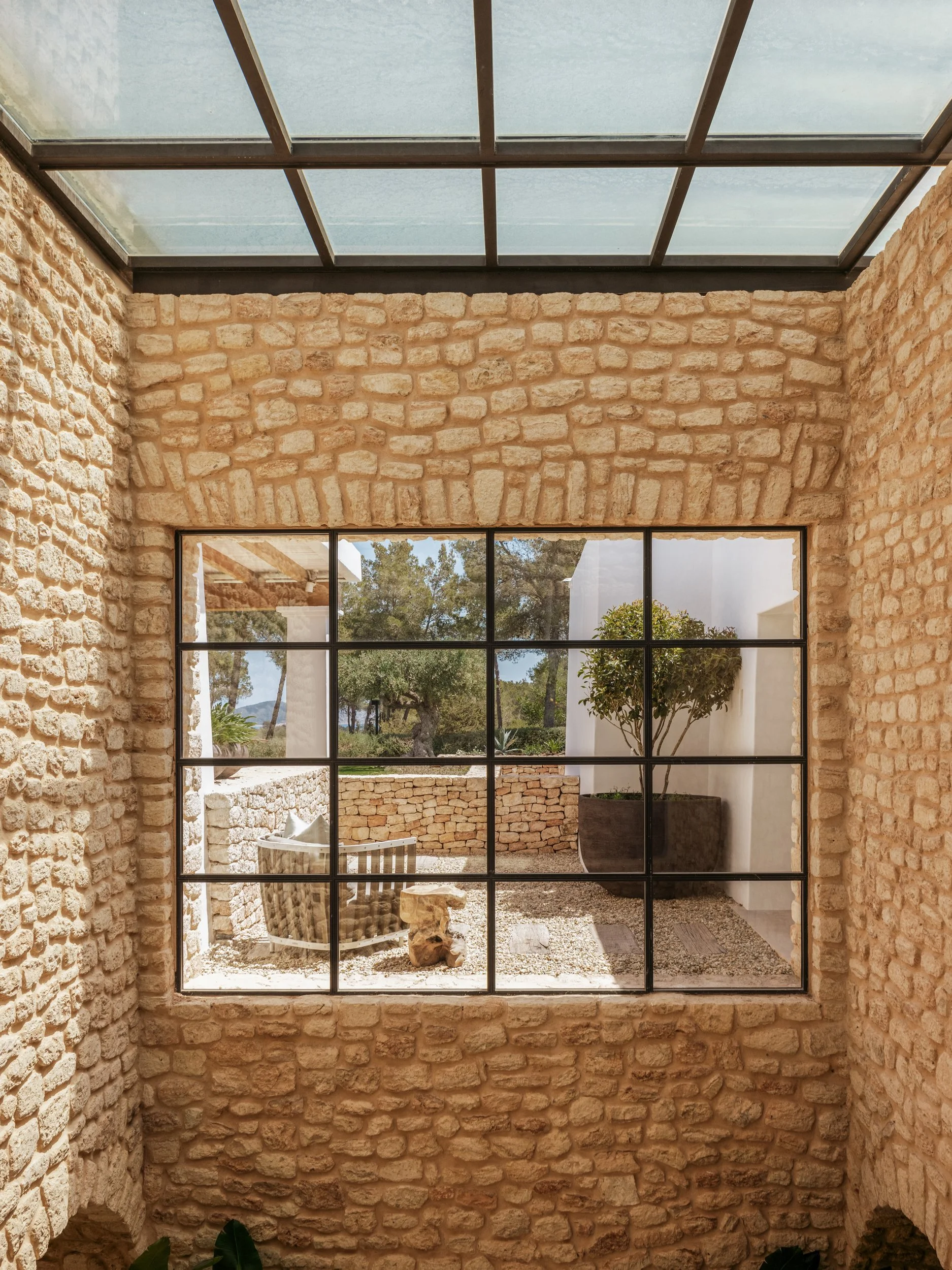 View through a large grid window showing an outdoor patio with stone flooring, a potted tree, a stone bench, and a decorative stone sculpture, surrounded by stone walls and trees in the background.
