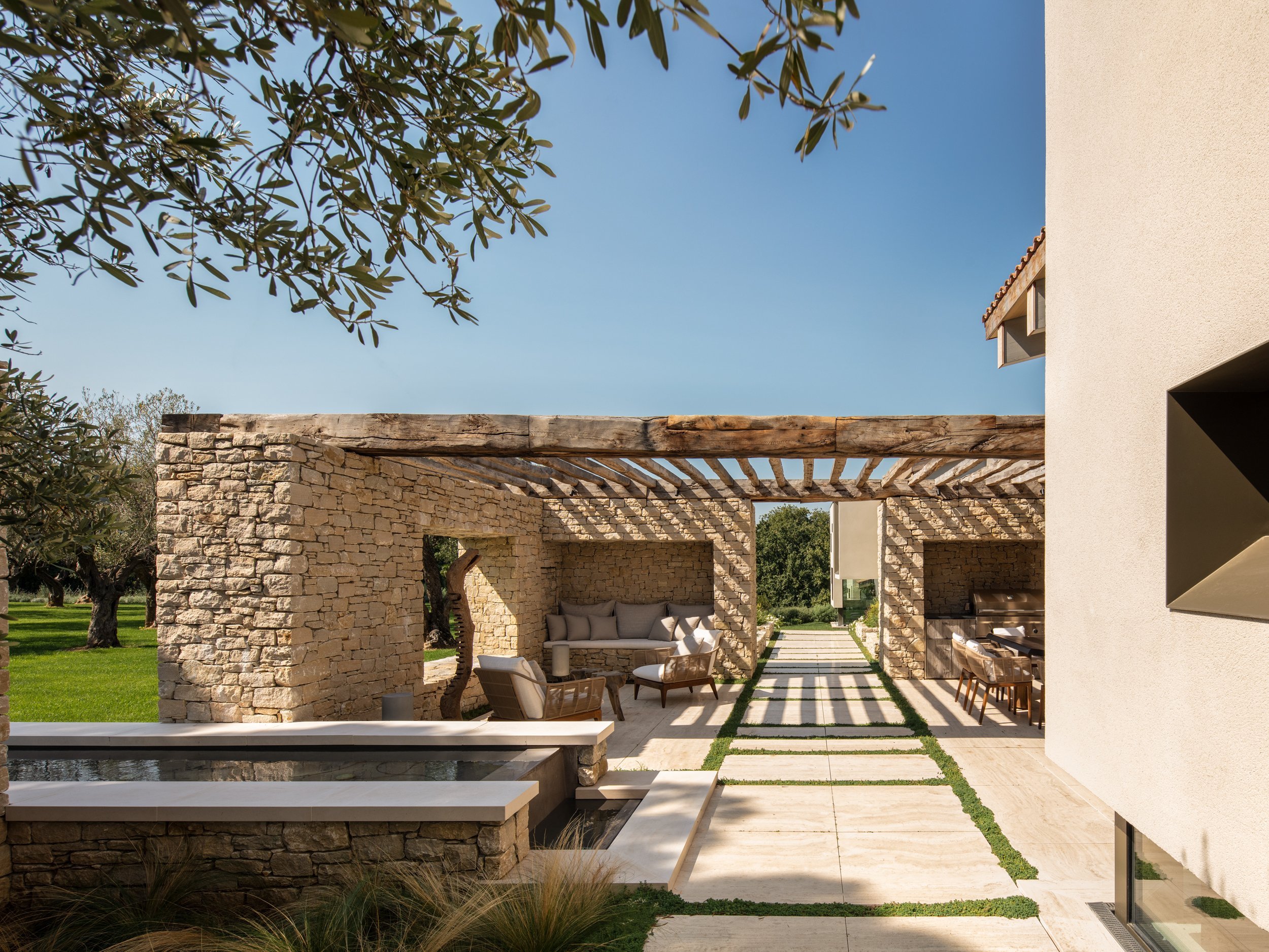 Modern outdoor patio with stone and wood architectural elements, sitting area with beige cushions, and a built-in grill, surrounded by greenery under a clear blue sky.