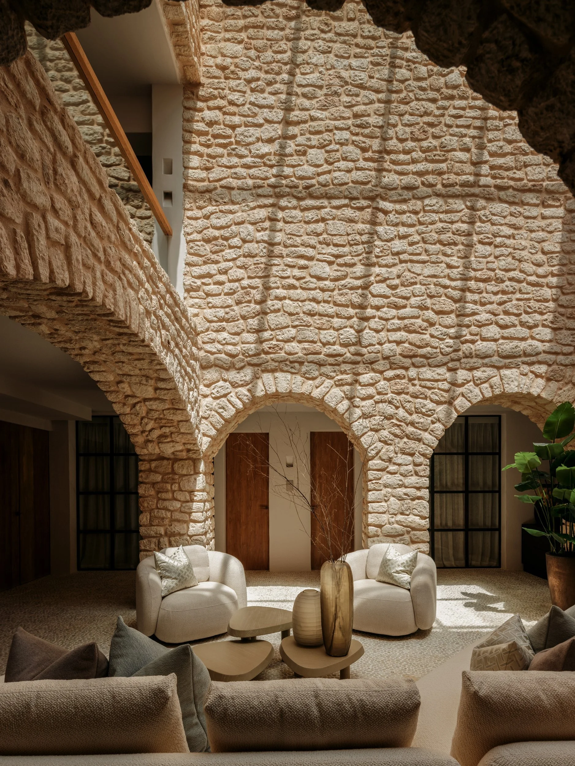 A modern living room with a rustic stone wall featuring arches and a curved balcony, furnished with white armchairs, a beige sofa, and decorative vases on a wooden table, with a sliding door and large window with curtains.