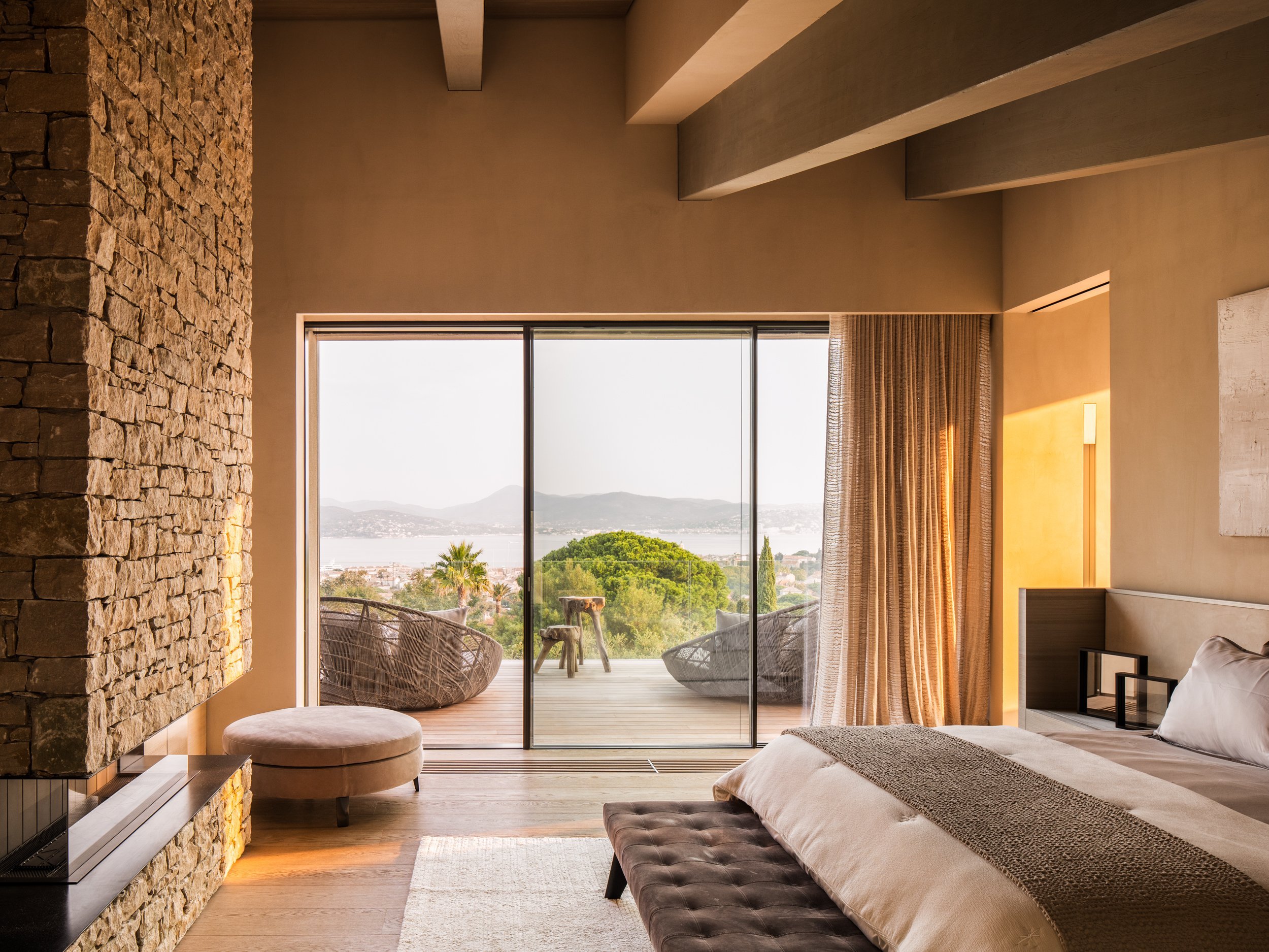 Modern bedroom with large glass sliding door, view of outdoor balcony with chairs, mountains, and water in the distance.