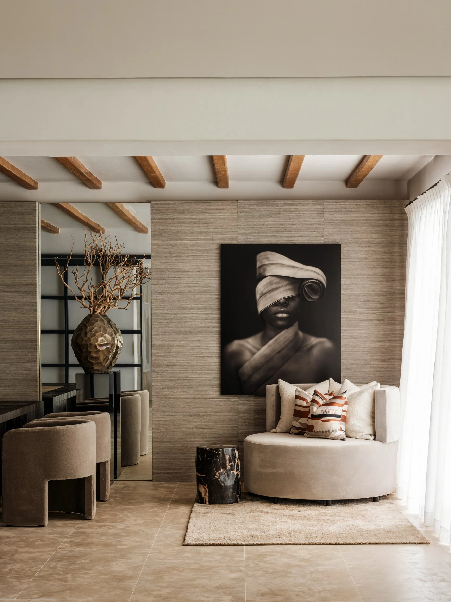 Modern living room with beige walls, wooden ceiling beams, black-and-white portrait art, round beige chair with striped pillows, a black marble side table, and decorative vase with twigs.
