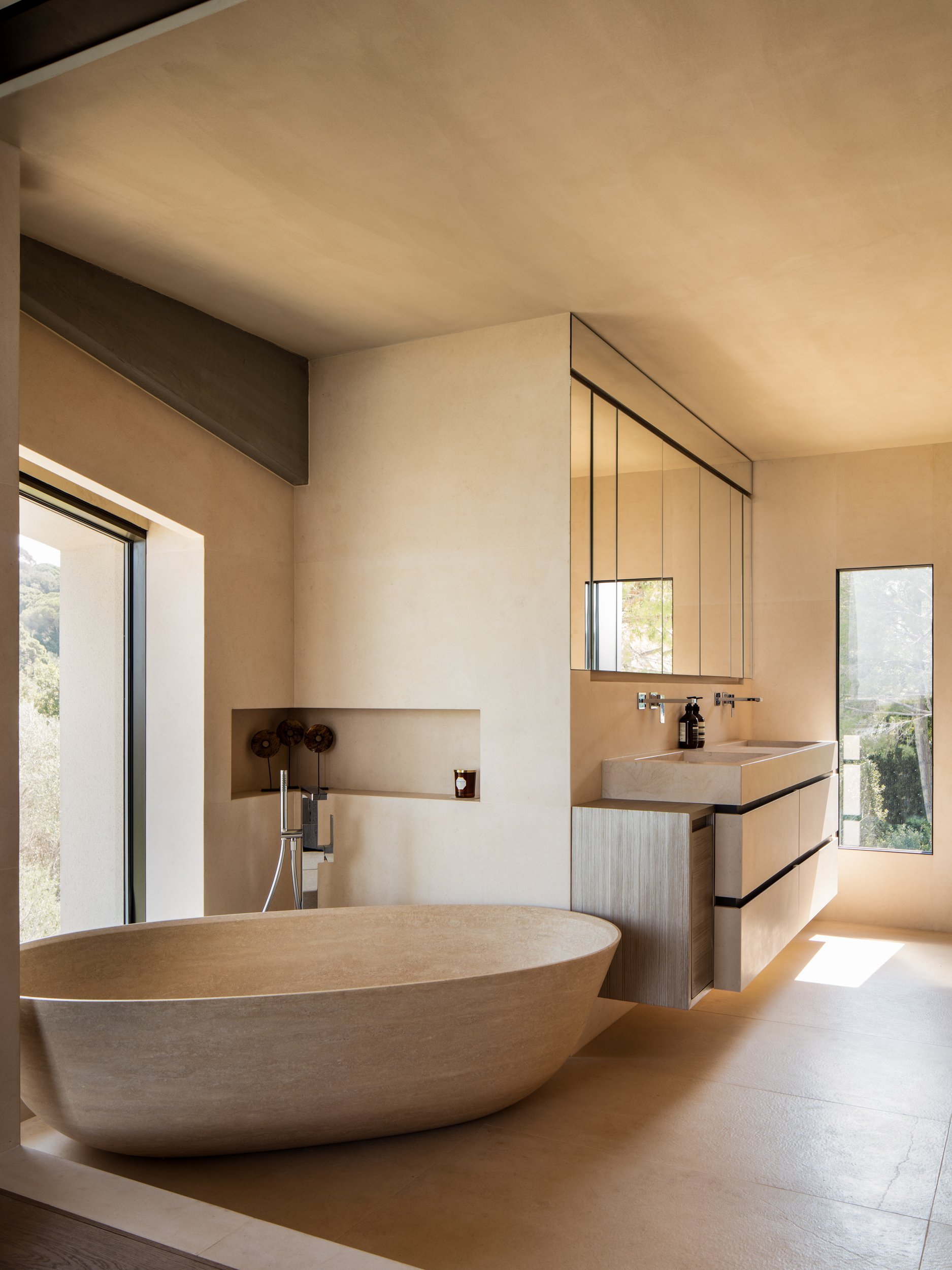 Modern minimalist bathroom with a freestanding oval bathtub, a wall-mounted sink, and a large mirror with black framing, illuminated by sunlight from large windows.