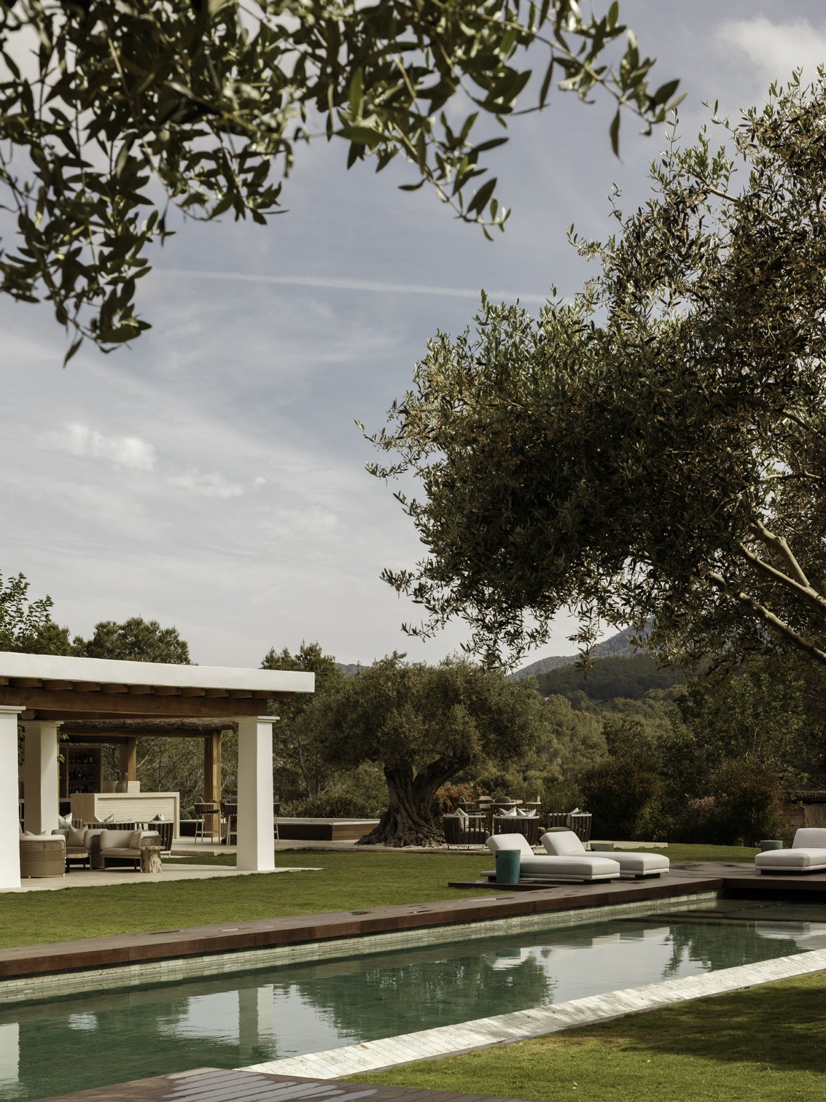 Outdoor poolside area with lounge chairs, a shaded seating area, trees, and a scenic background of hills under a partly cloudy sky.