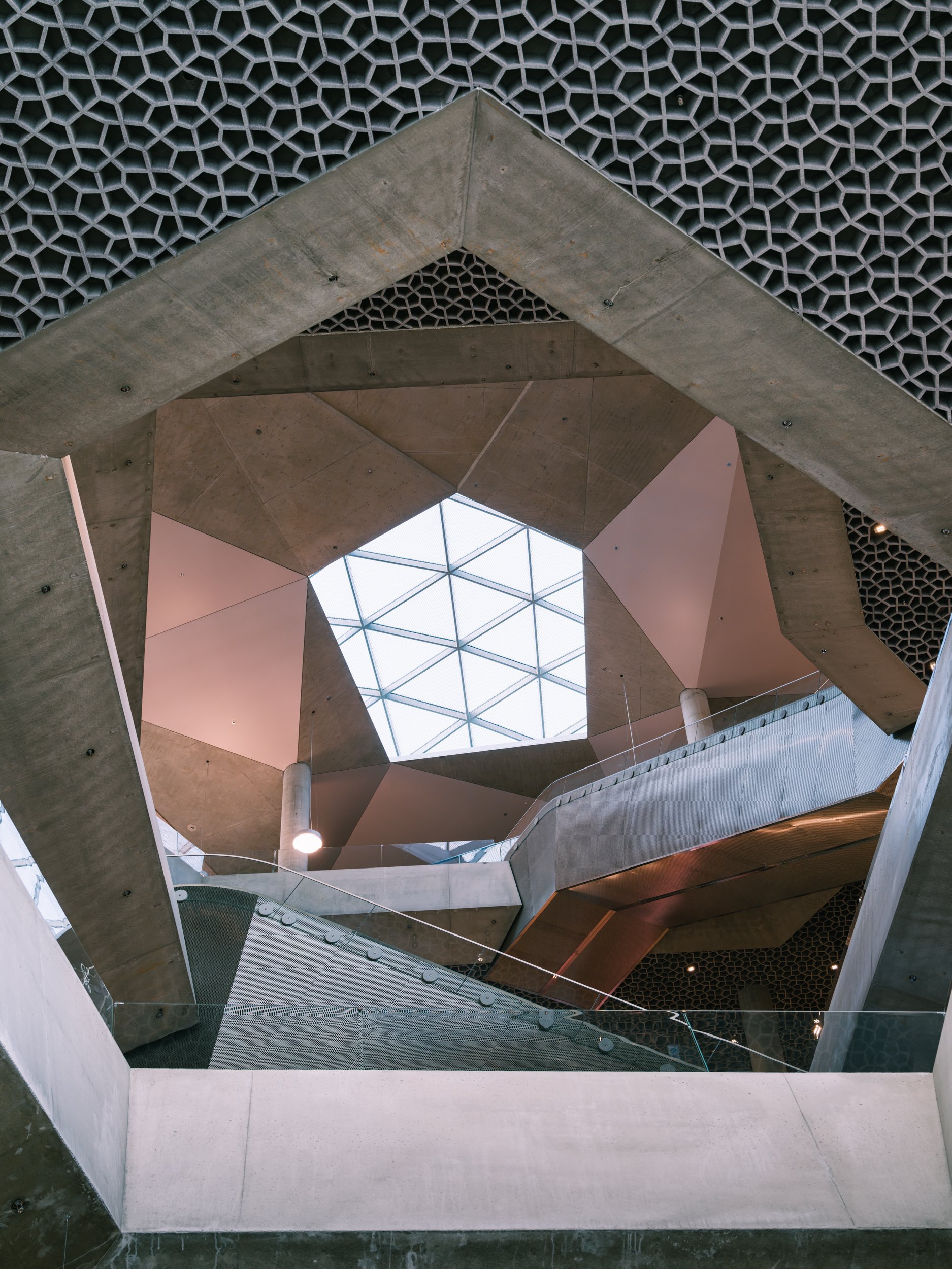 Interior view of a modern building with geometric concrete structures, glass ceiling, and intricate honeycomb-patterned panels.