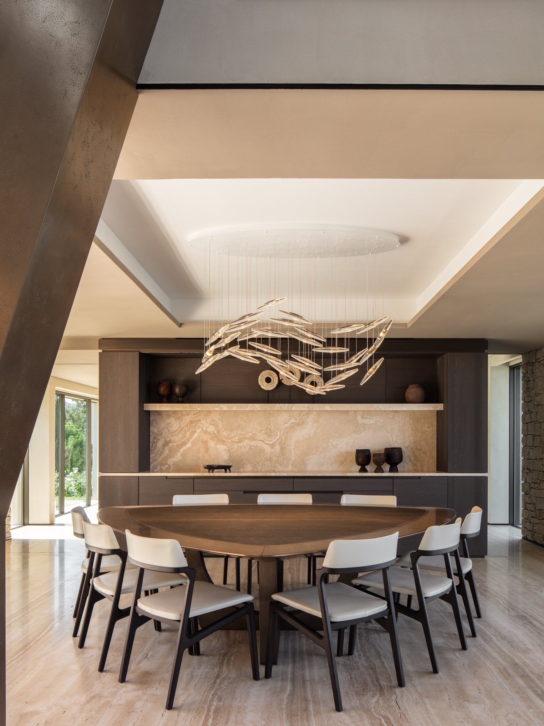 Modern dining room with a round wooden table, white chairs, a decorative light fixture hanging from the ceiling, and a wall with dark cabinetry and a beige marble backsplash.