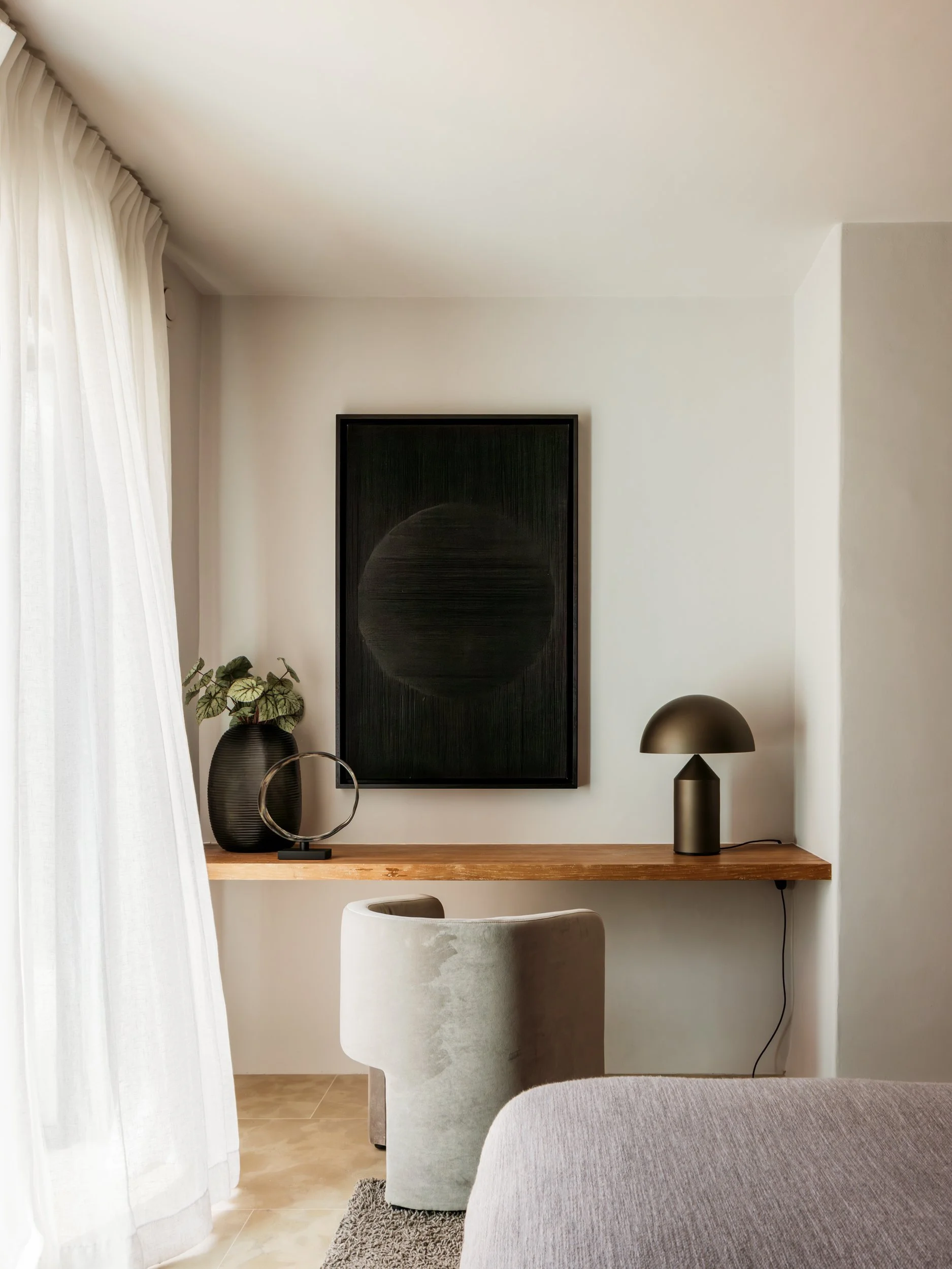 A minimalist living room corner with a wooden desk, a green velvet chair, a black vase with green plants, a black circular sculpture, and a black modern table lamp on the desk. A large abstract black art piece hangs on the white wall, and sheer white