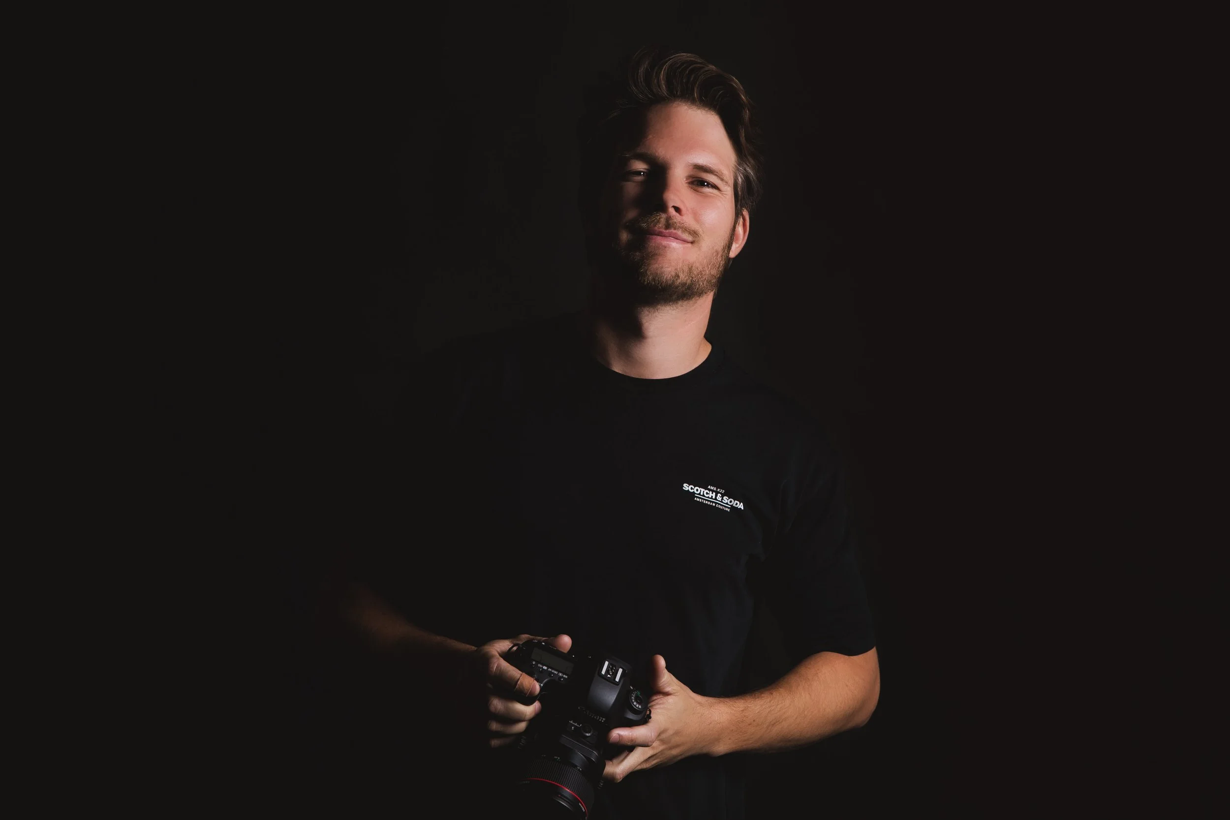 A young man with light brown hair and a beard, wearing a black t-shirt with a small white logo that says "Scotch & Soda," holding a professional camera in his hands, standing against a dark background.