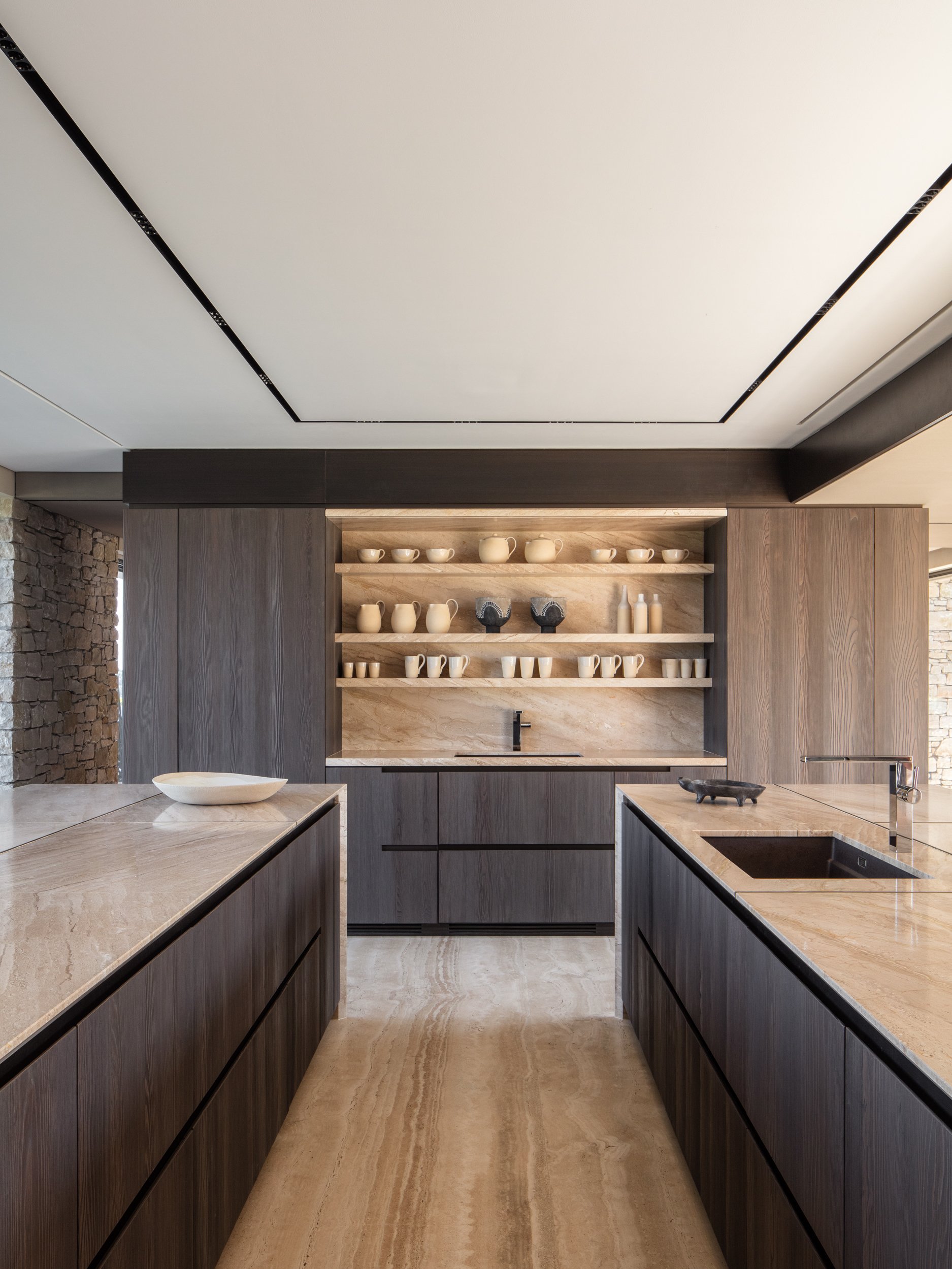 Modern kitchen with beige marble countertops, dark wooden cabinets, open shelves with ceramics, stone accents, and a sink.