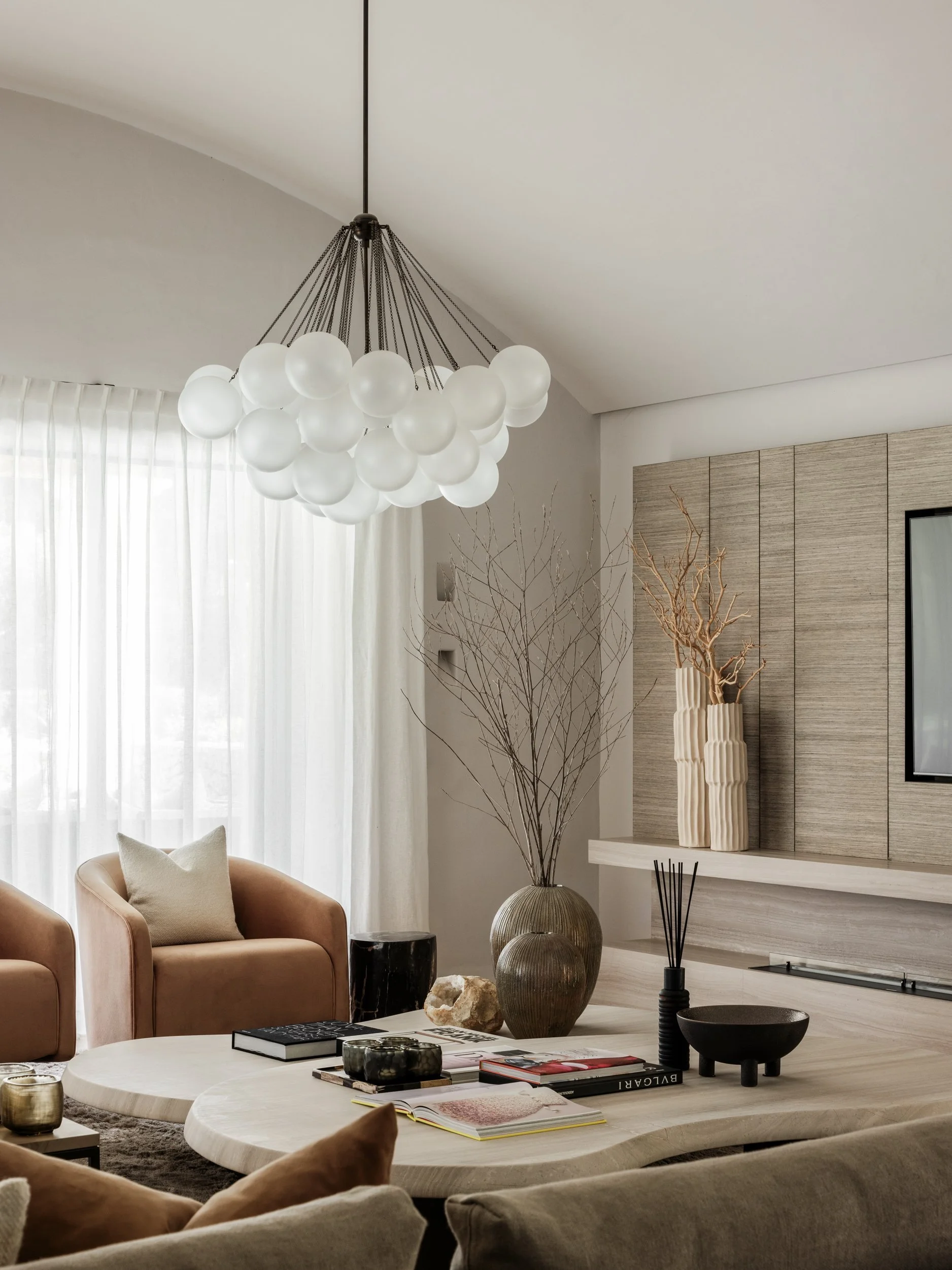 Modern living room with a round, white coffee table, beige armchairs, decorative vases, a large leafy plant, and a contemporary chandelier hanging from the ceiling.