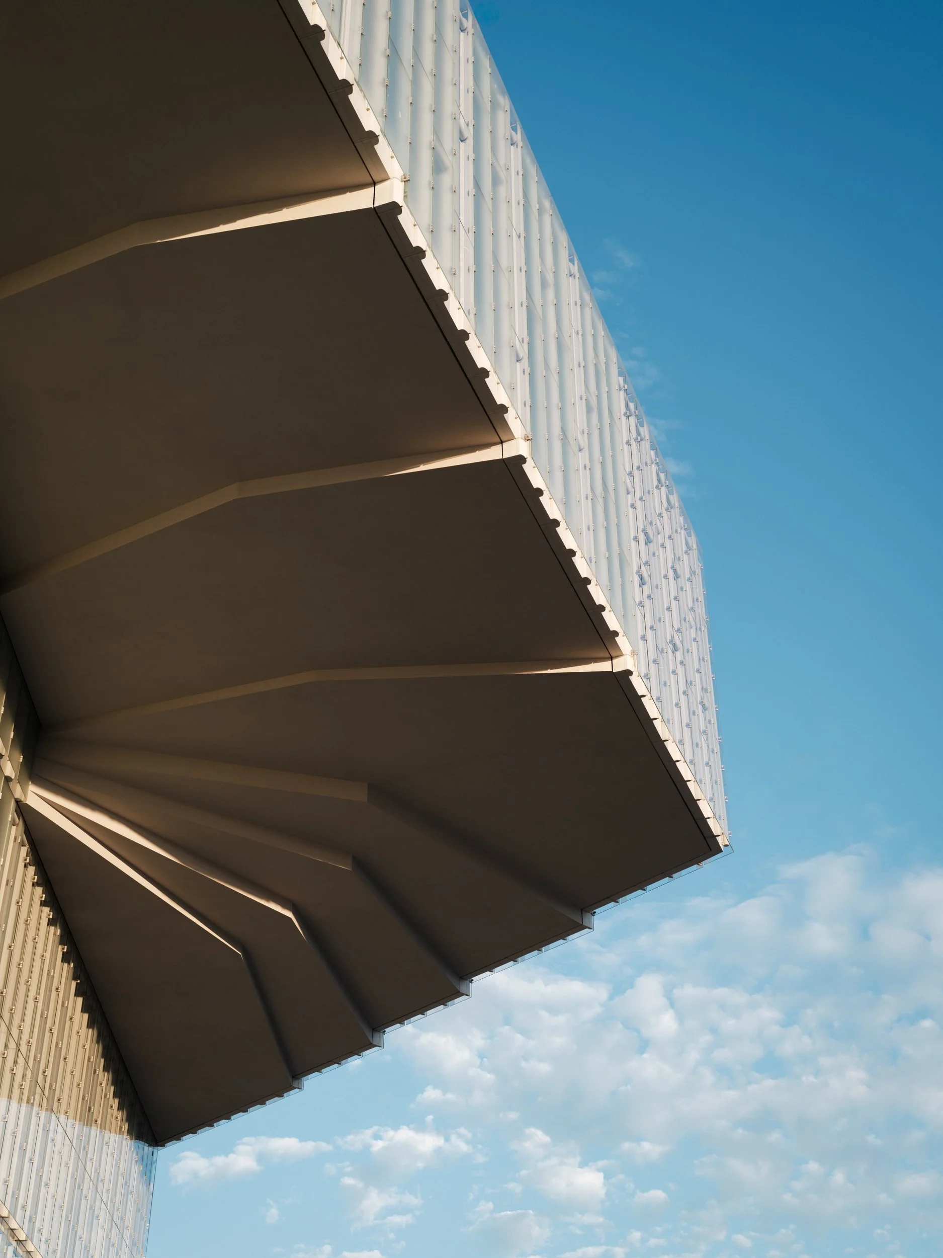 Close-up of modern architectural building with overhanging roof, set against a partly cloudy blue sky.