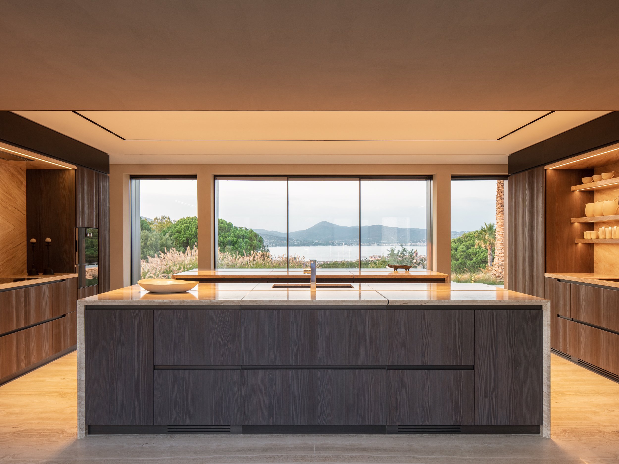 Modern kitchen with central island, large window view of trees, mountains, and water in the background, and built-in open shelving on right with plates and bowls.
