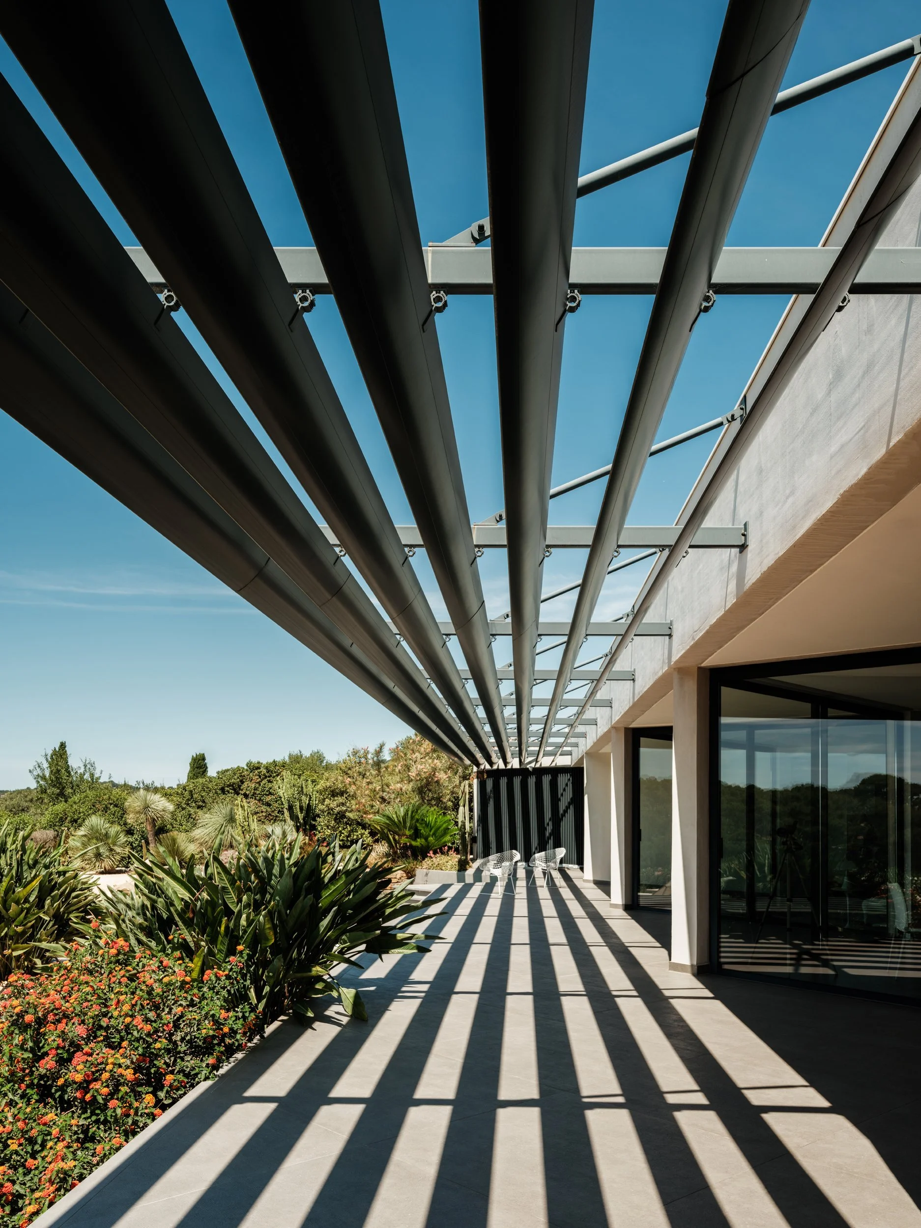 Modern house with a patio covered by a structure with metal beams casting striped shadows, surrounded by desert plants and greenery, under a clear blue sky.