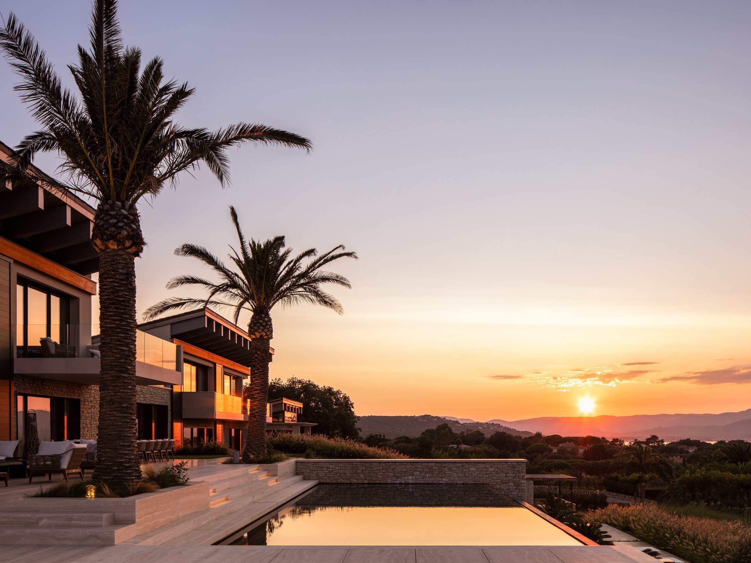 Luxury modern house with palm trees at sunset, overlooking a landscape with mountains, some outdoor seating, and a swimming pool reflecting the sky.