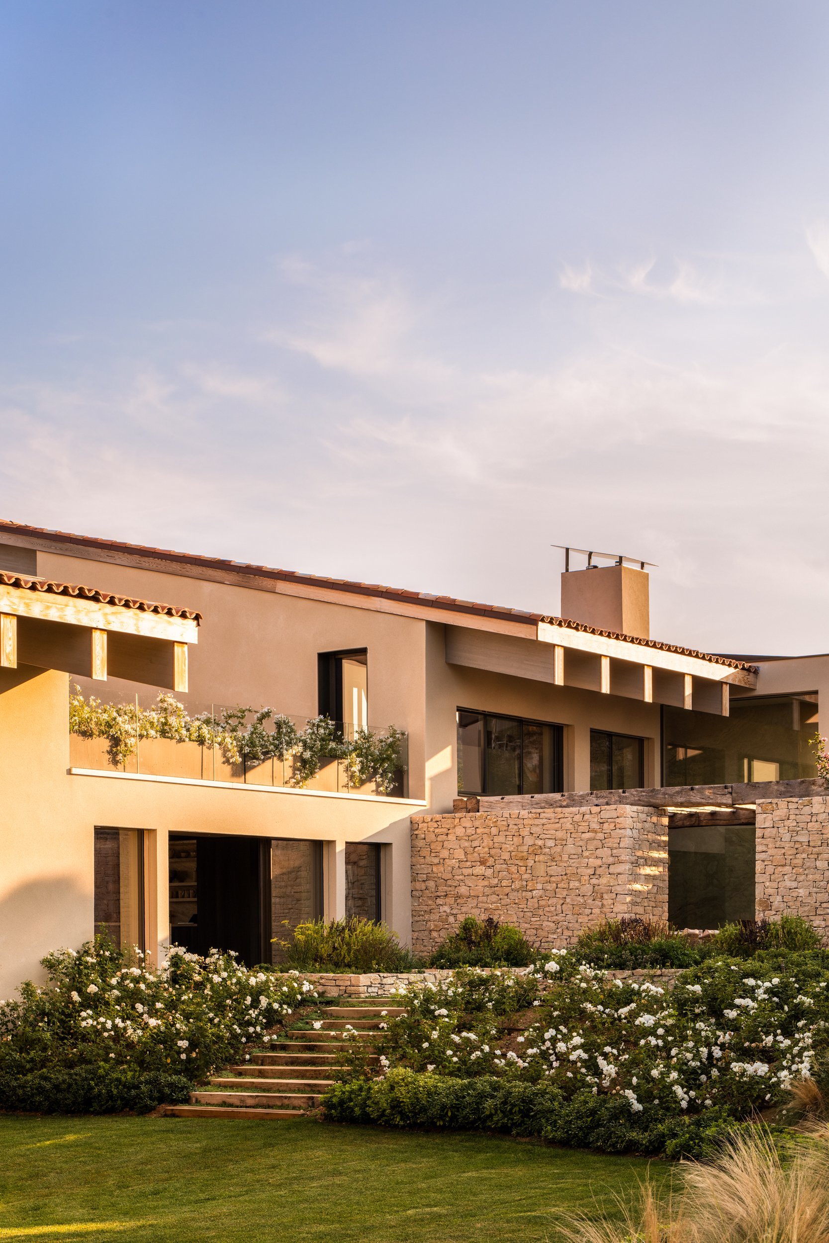 A modern house with beige walls, glass doors, stone accents, and a landscaped garden with white flowers and green shrubbery, under a clear sky.