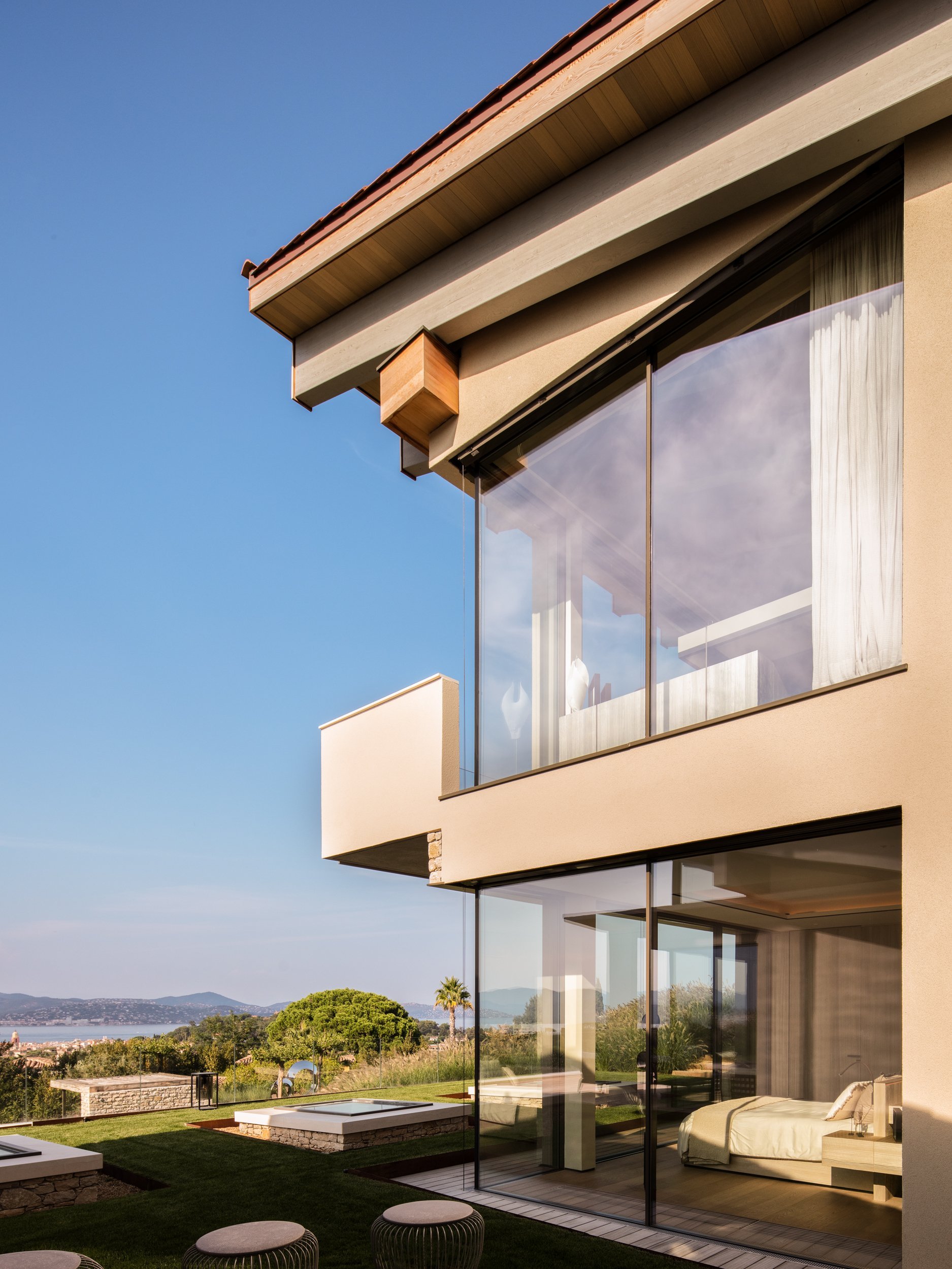 Close-up of a modern house with large glass windows, beige walls, and a wooden overhang, overlooking a scenic landscape with hills, trees, and a bright blue sky.