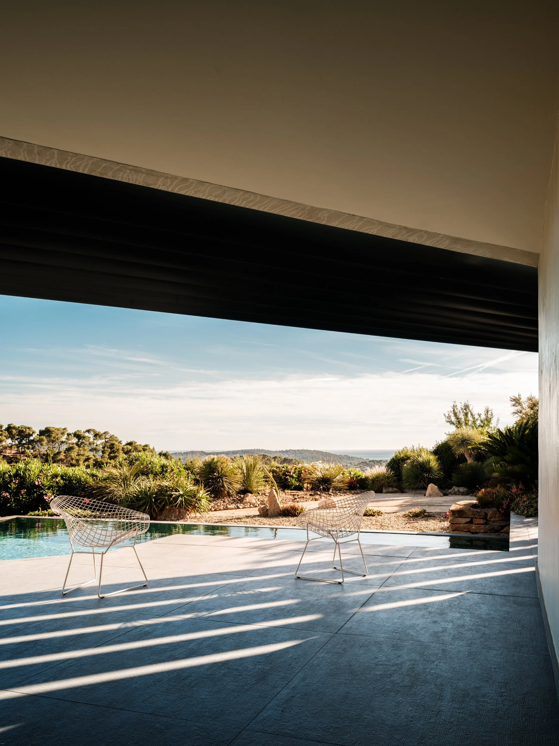 Modern outdoor patio with two white metal chairs overlooking a lush landscape with desert plants and a clear sky in the distance.