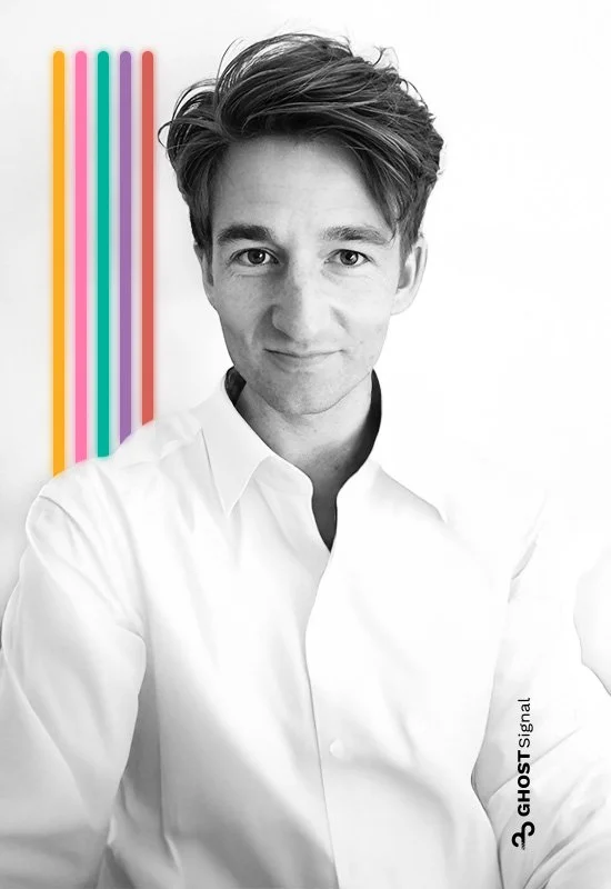 A young man with styled brown hair and light skin, wearing a white shirt, standing in front of white background with colorful vertical lines on the left.