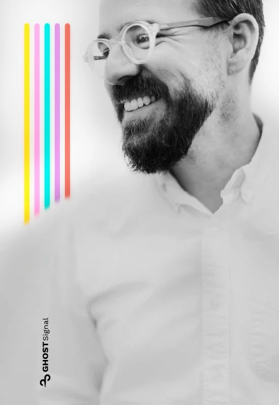 A smiling man with a beard and glasses, wearing a white shirt, standing in front of a white background with colorful vertical lines.