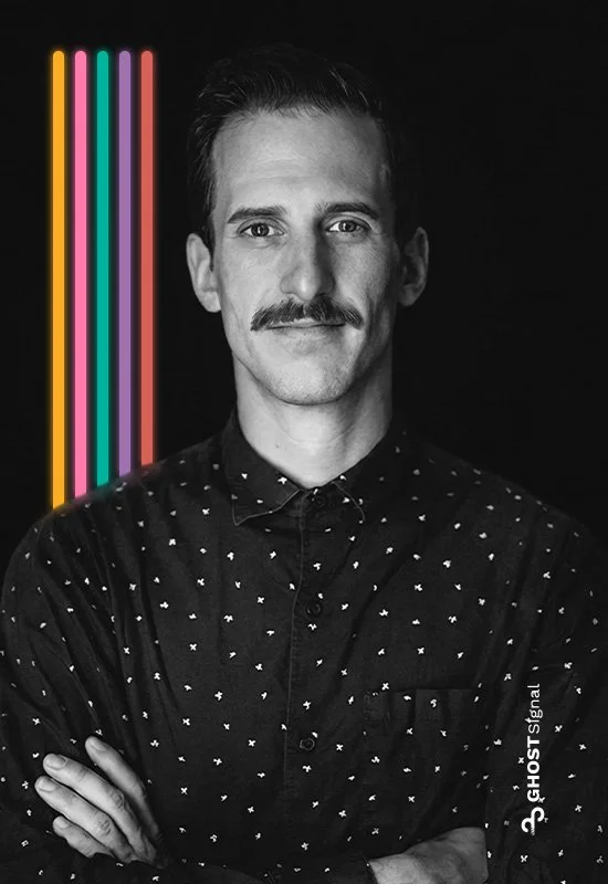 Black and white portrait of a man with a mustache, wearing a dotted shirt and crossing his arms. Neon vertical lines in pink, yellow, blue, and green are on the left side of the image.