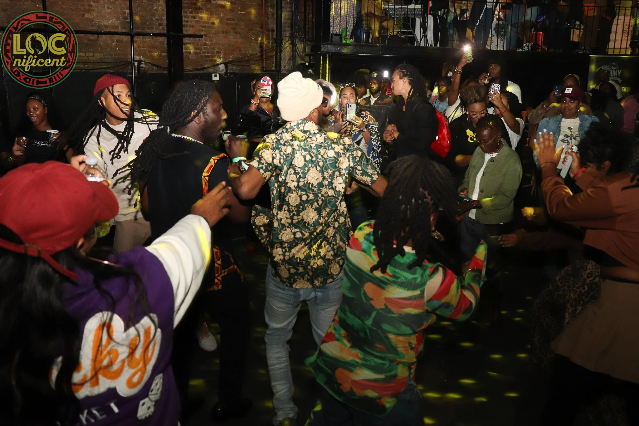 A diverse group of people, mostly young adults, dancing and enjoying music at an indoor party or club. The background features a brick wall and a dancing crowd with colorful lighting and some people taking pictures with their phones.