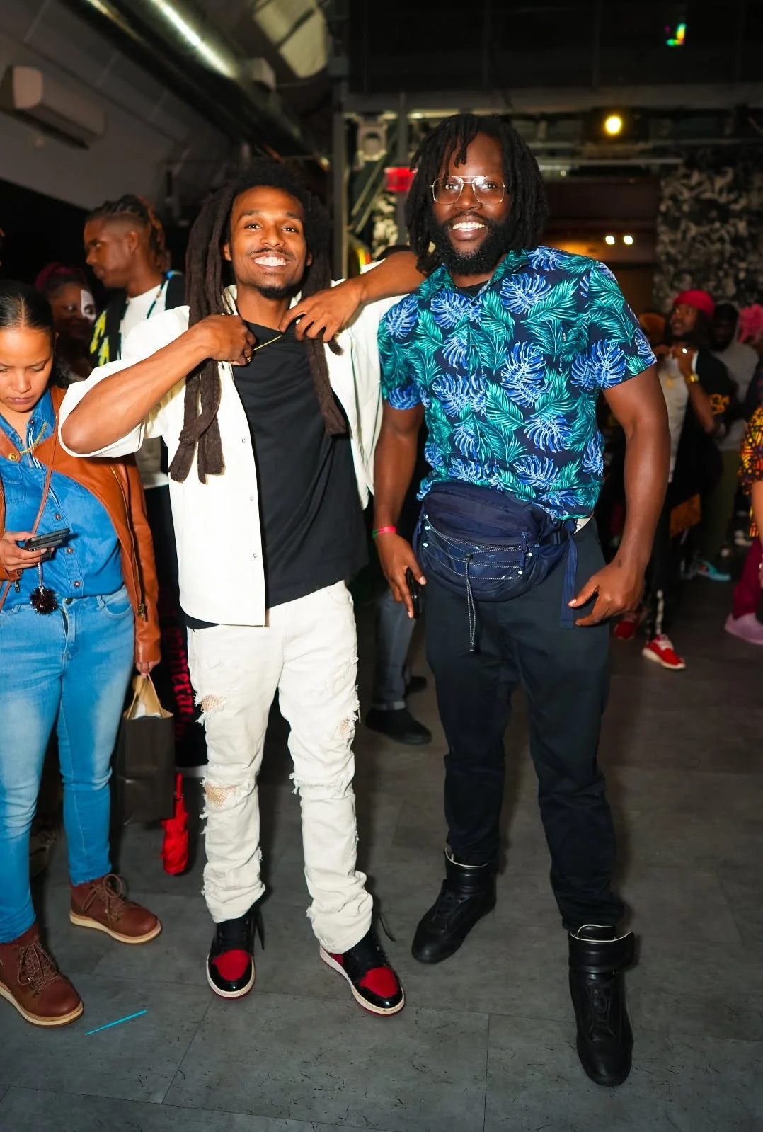 Two men smiling and posing together at an indoor event, surrounded by other people.