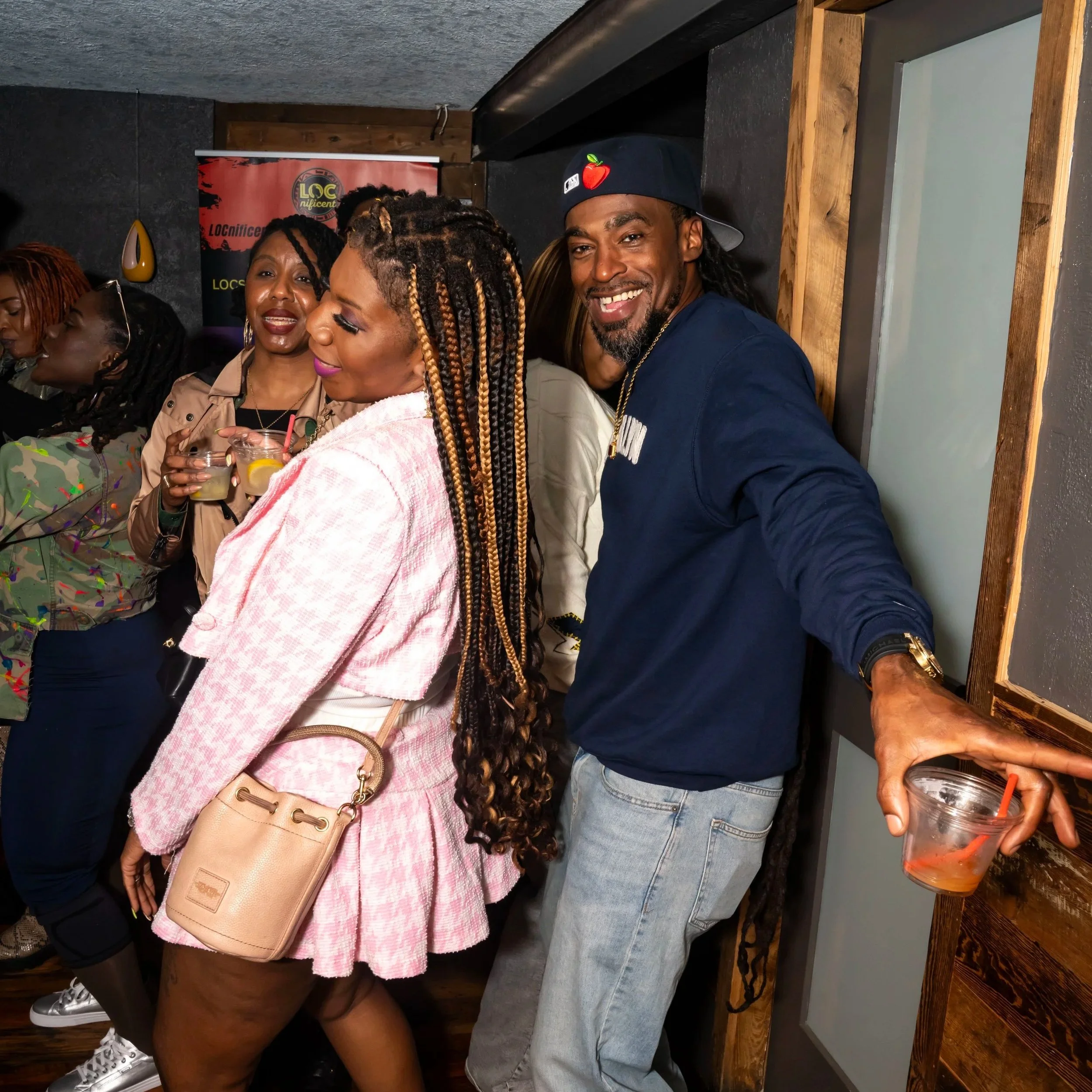 A group of diverse people gathered together at a social event, smiling, dancing, and enjoying drinks in a dimly lit indoor setting.