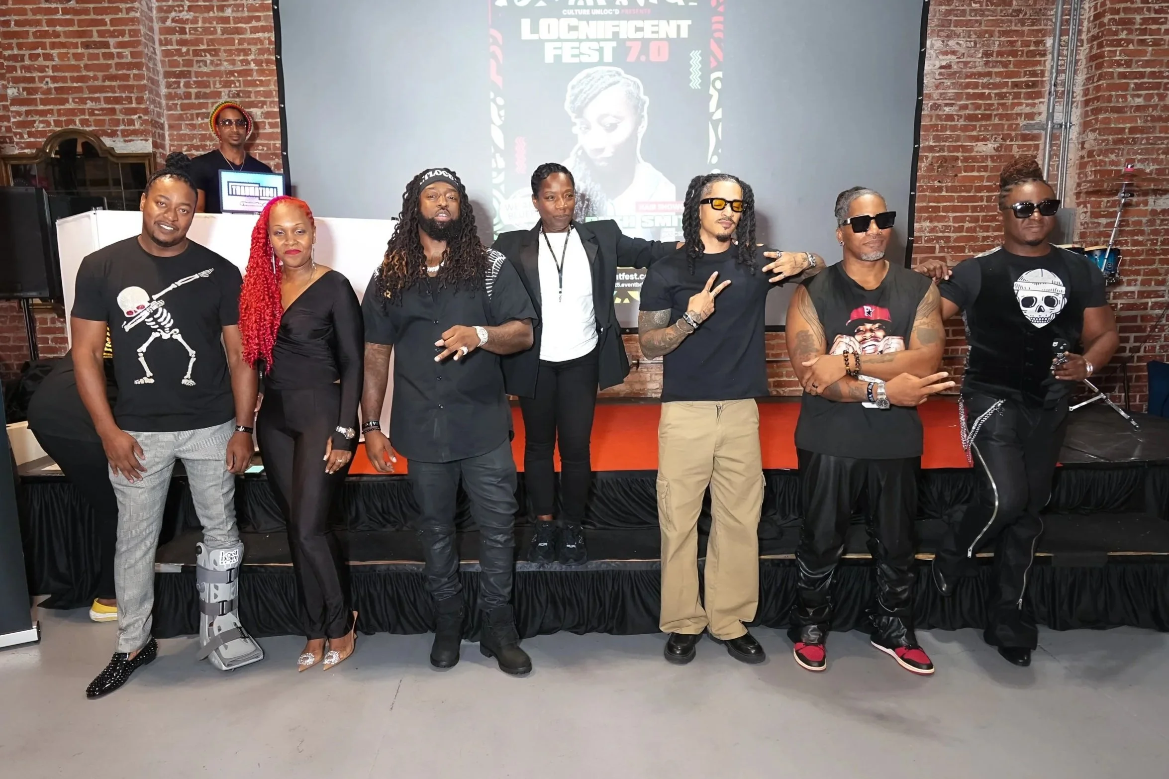 Group of eight people standing in front of a promotional display for Locnificent Fest 7.0, with a brick wall background, at an indoor event.