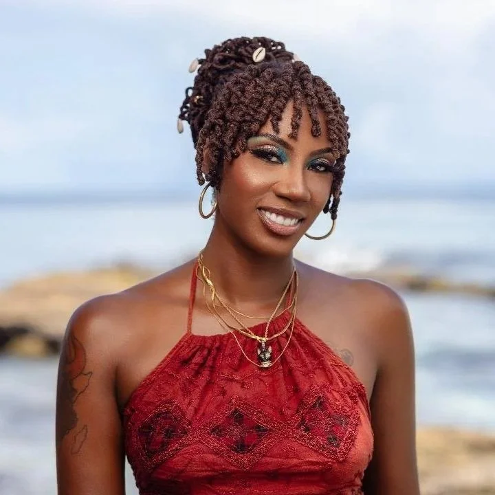 A woman with dark skin and short, curly, textured hair decorated with seashells, standing on the beach with ocean and rocks in the background, wearing a red halter top, gold jewelry, and makeup.