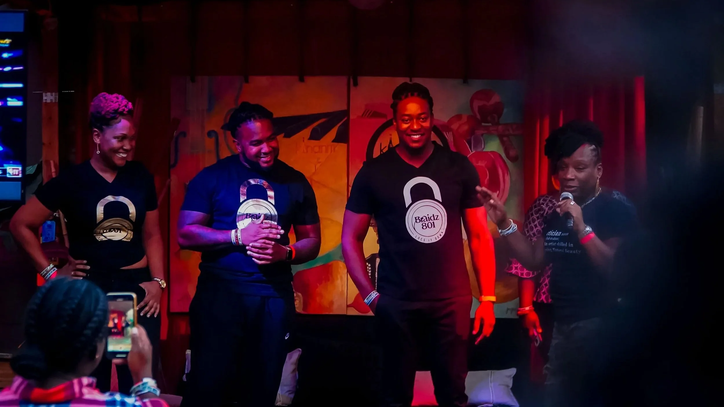 Four people, three women and one man, are standing on a stage at an event. They are wearing black t-shirts with a gold and white logo that says 'Braidz 801' and have microphones. The background is colorful with a mural, and there are people in the audience taking pictures.