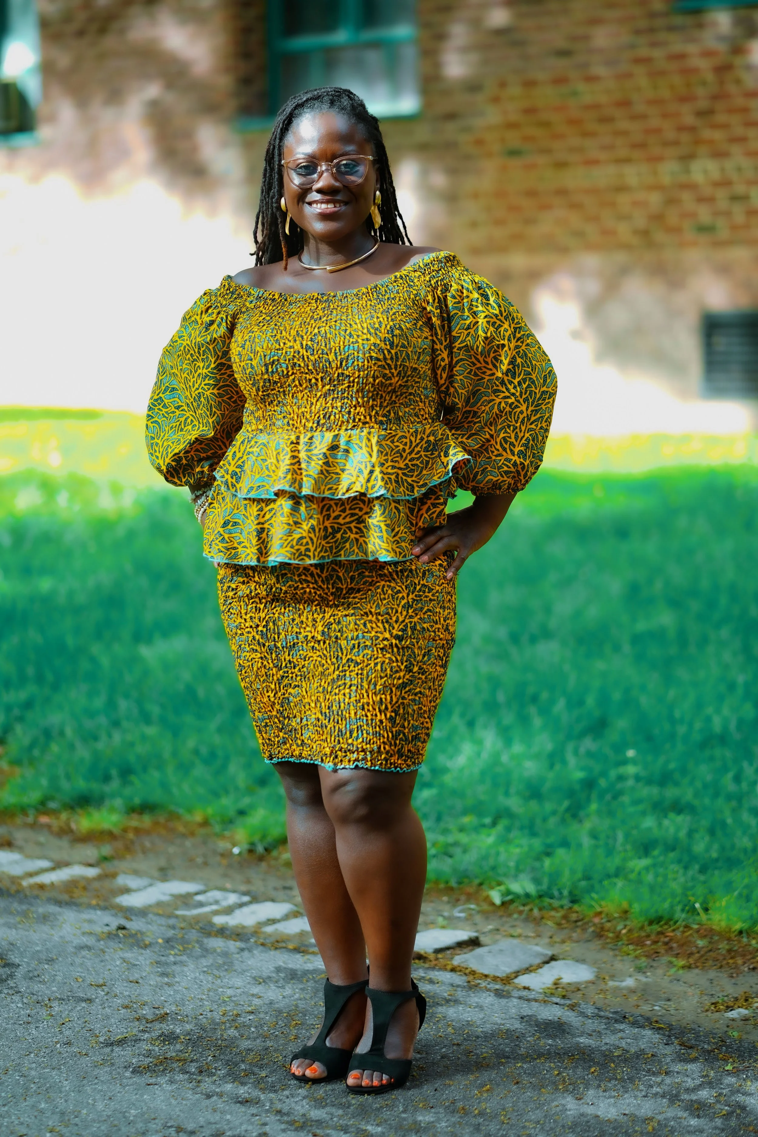 A woman wearing glasses and traditional African print clothing, standing outdoors on a paved path with green grass in the background.