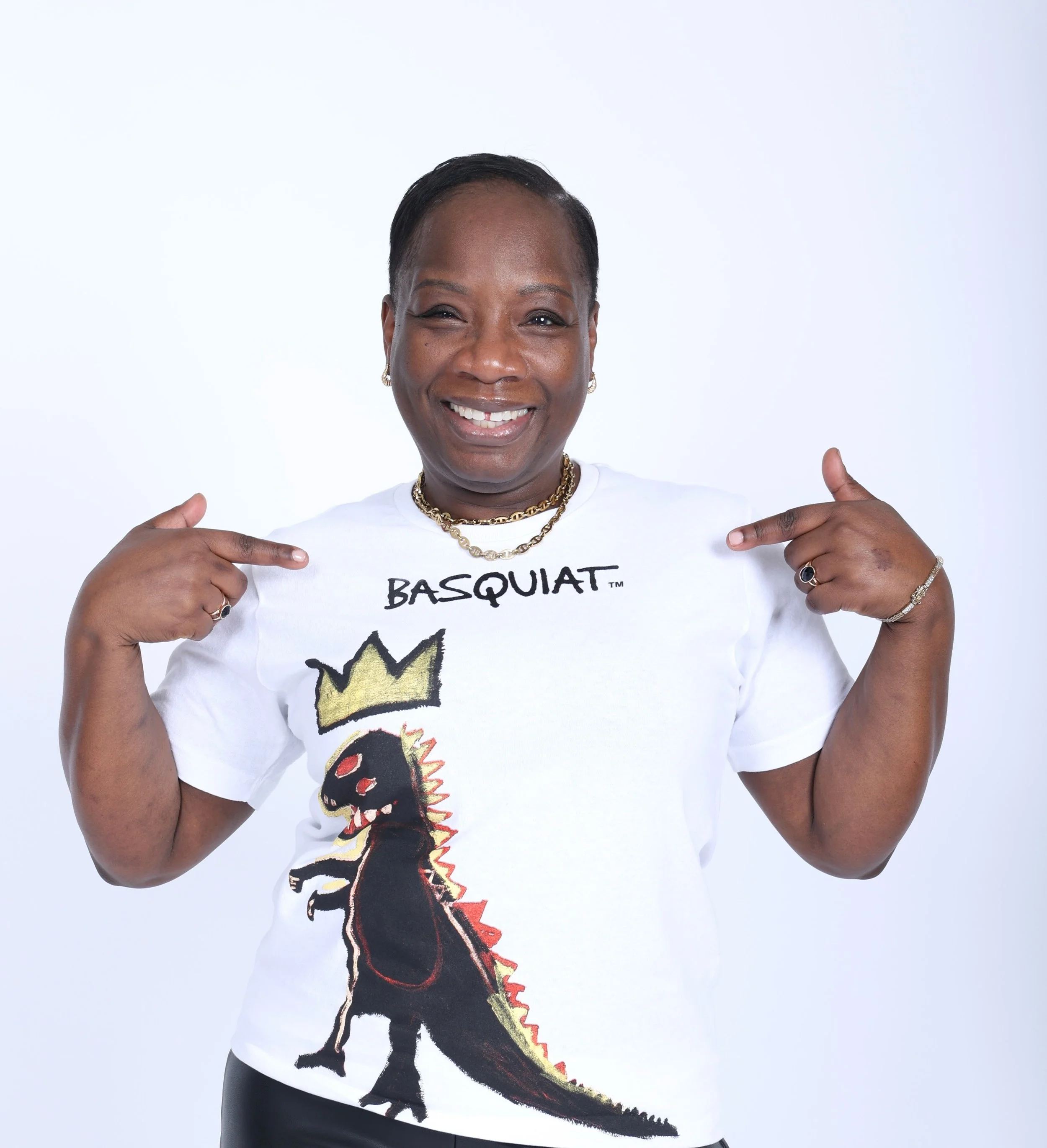 Woman smiling, pointing at her t-shirt with a dinosaur graphic and the word 'BASQUIAT' printed on it, against a plain white background.