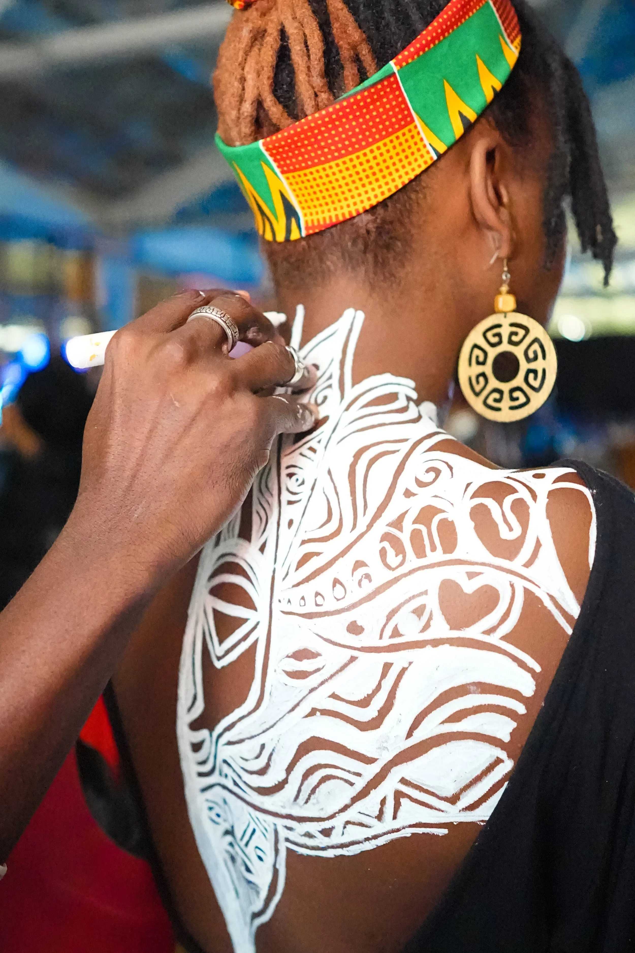 A woman is wearing a colorful headwrap, large circular earrings, and a white lace top with intricate patterns. She is painting or drawing white designs on her back with a small brush or tool. Her skin is dark, and she is inside a place with vibrant, blurred background lighting.