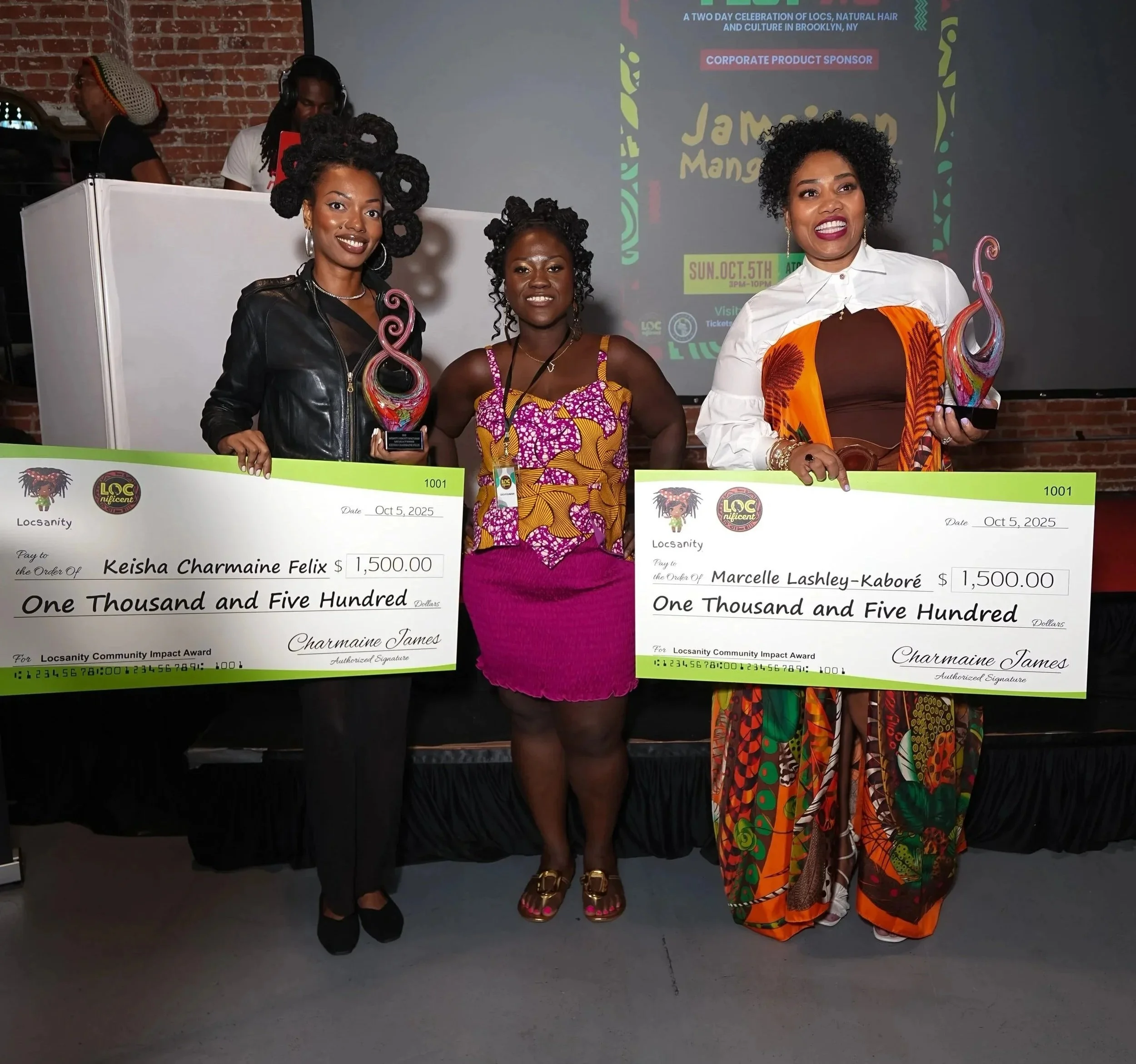 Three women standing with large ceremonial checks at an event. The woman on the left holds a colorful trophy, wears a black leather jacket, and has styled black hair. The woman in the middle wears a vibrant pink, yellow, and purple patterned dress, and the woman on the right wears a white shirt with an orange and patterned fabric underneath. They are smiling in front of a brick wall and a screen displaying event information.