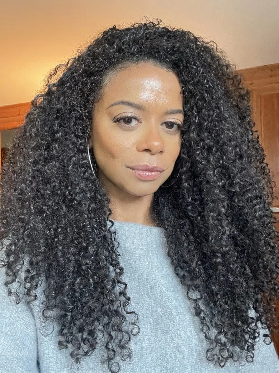 A woman with curly black hair, wearing a light grey sweater and hoop earrings, is indoors with wooden furniture and a beige wall behind her.