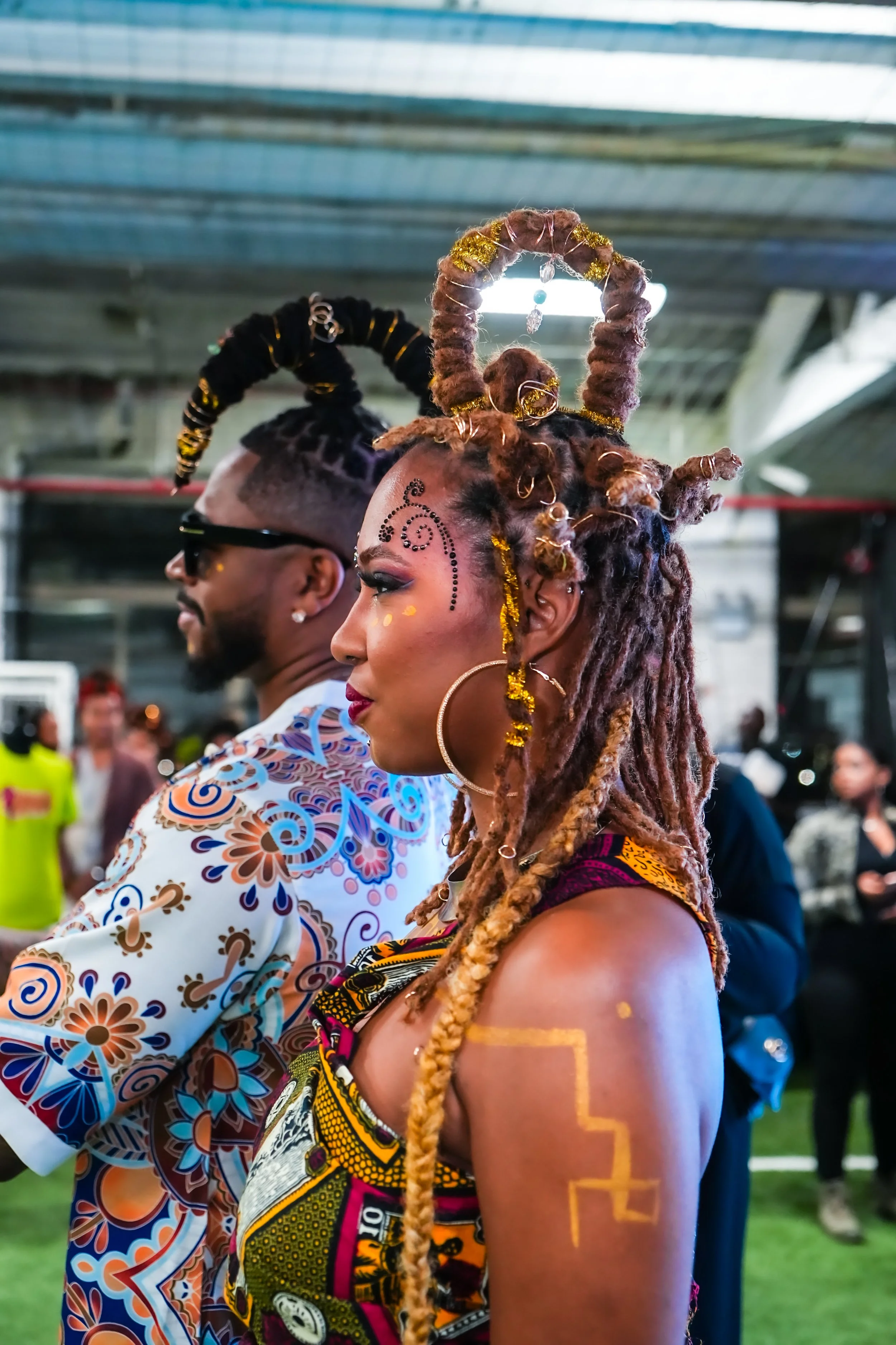 Two individuals dressed in vibrant traditional African clothing with elaborate head wraps and face decorations, standing side by side at an outdoor event.