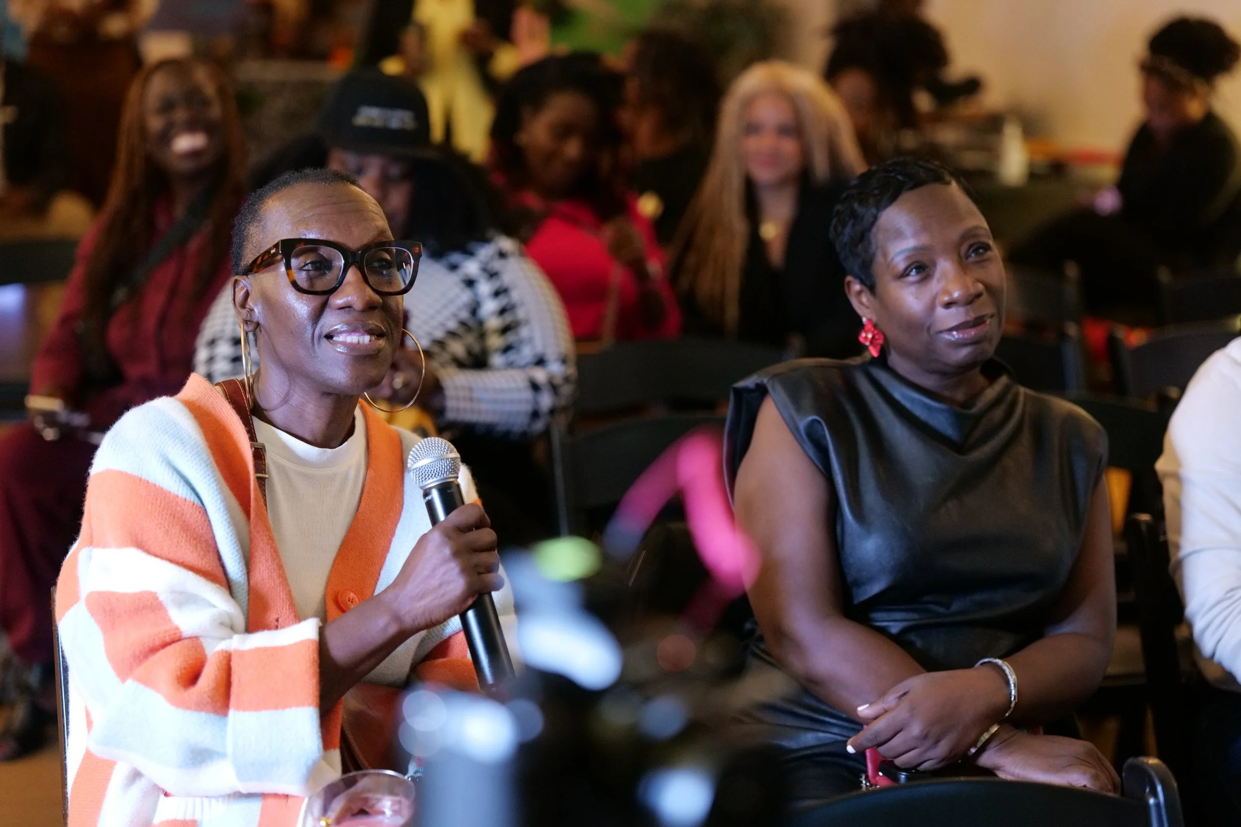 A diverse group of women attending an indoor event, with some women smiling and one woman holding a microphone.