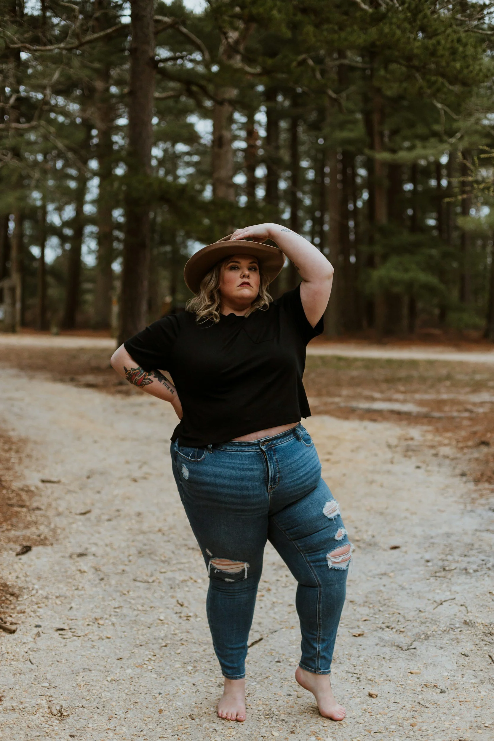 A woman with blonde hair, tattoos, and a serious expression, standing on a dirt path in a forest, wearing a wide-brimmed hat, black t-shirt, and ripped jeans, with one hand on her hip and the other resting on her hat.